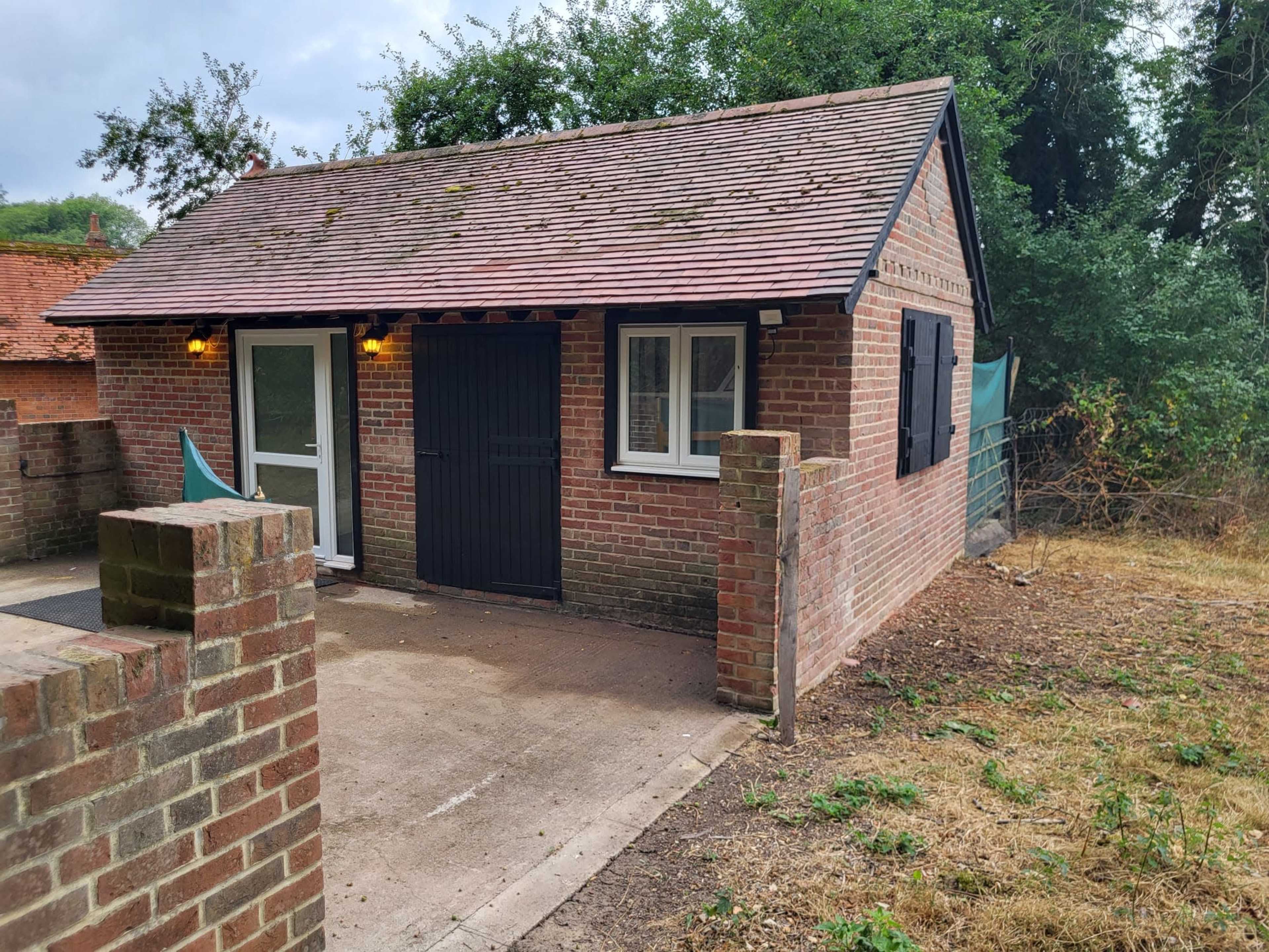 A small brick cottage with a black door, surrounded by a grassy area and trees.