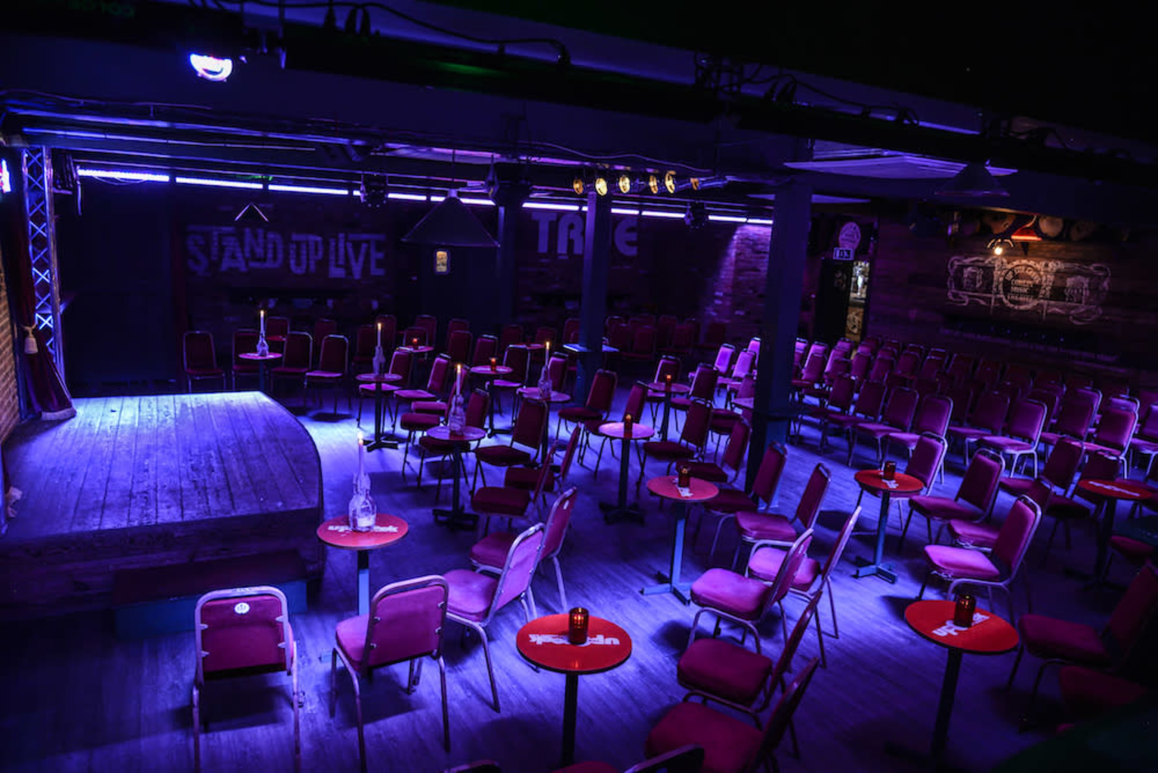 The image shows a dimly lit comedy club with red chairs and small round tables arranged in rows facing a stage.