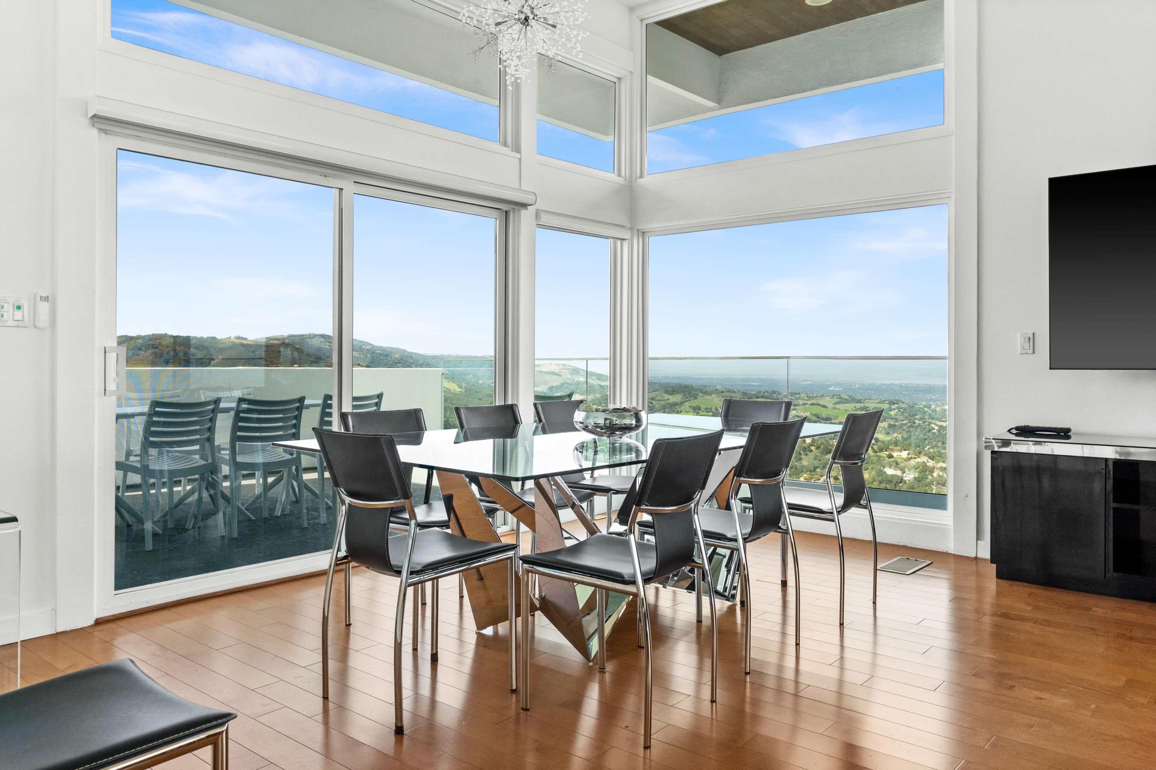 A modern dining room features a glass table surrounded by black chairs, large windows that offer a view of the landscape, and a chandelier hanging overhead.