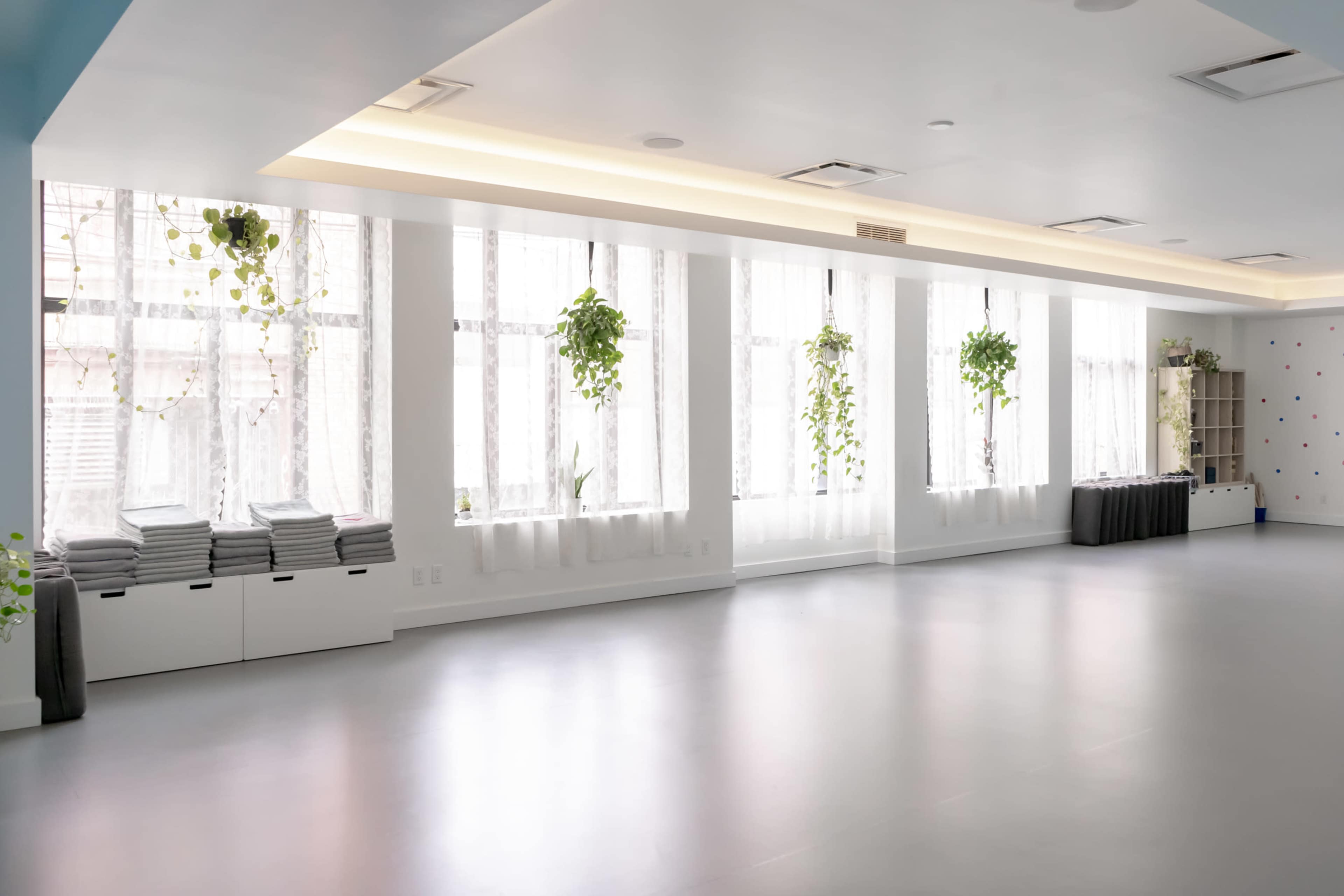 A bright, minimalist room features large windows with hanging plants, neatly stacked towels, and a shelving unit along the wall.