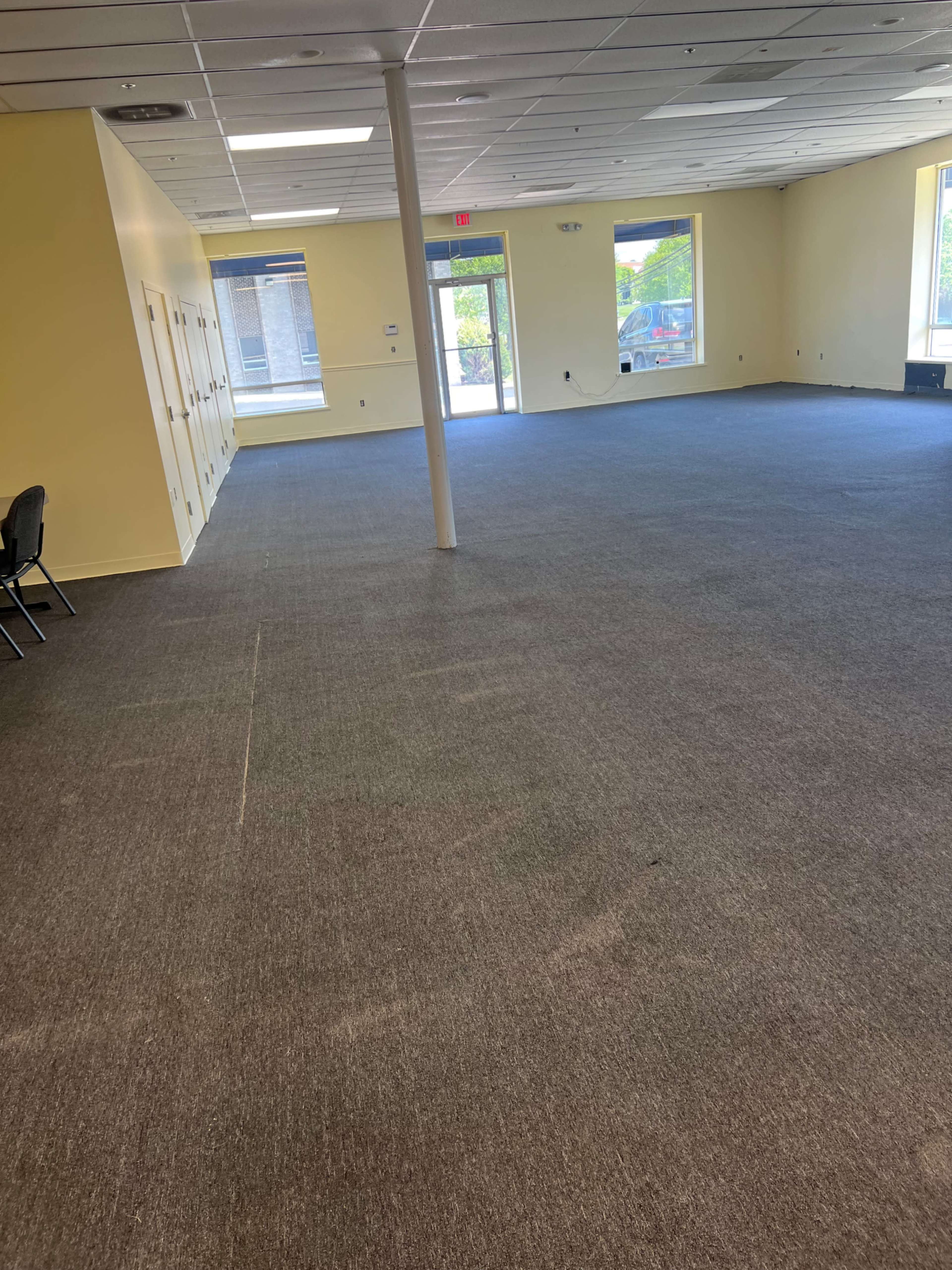 The image shows a spacious, empty room with yellow walls, gray carpeting, and large windows allowing natural light to enter.