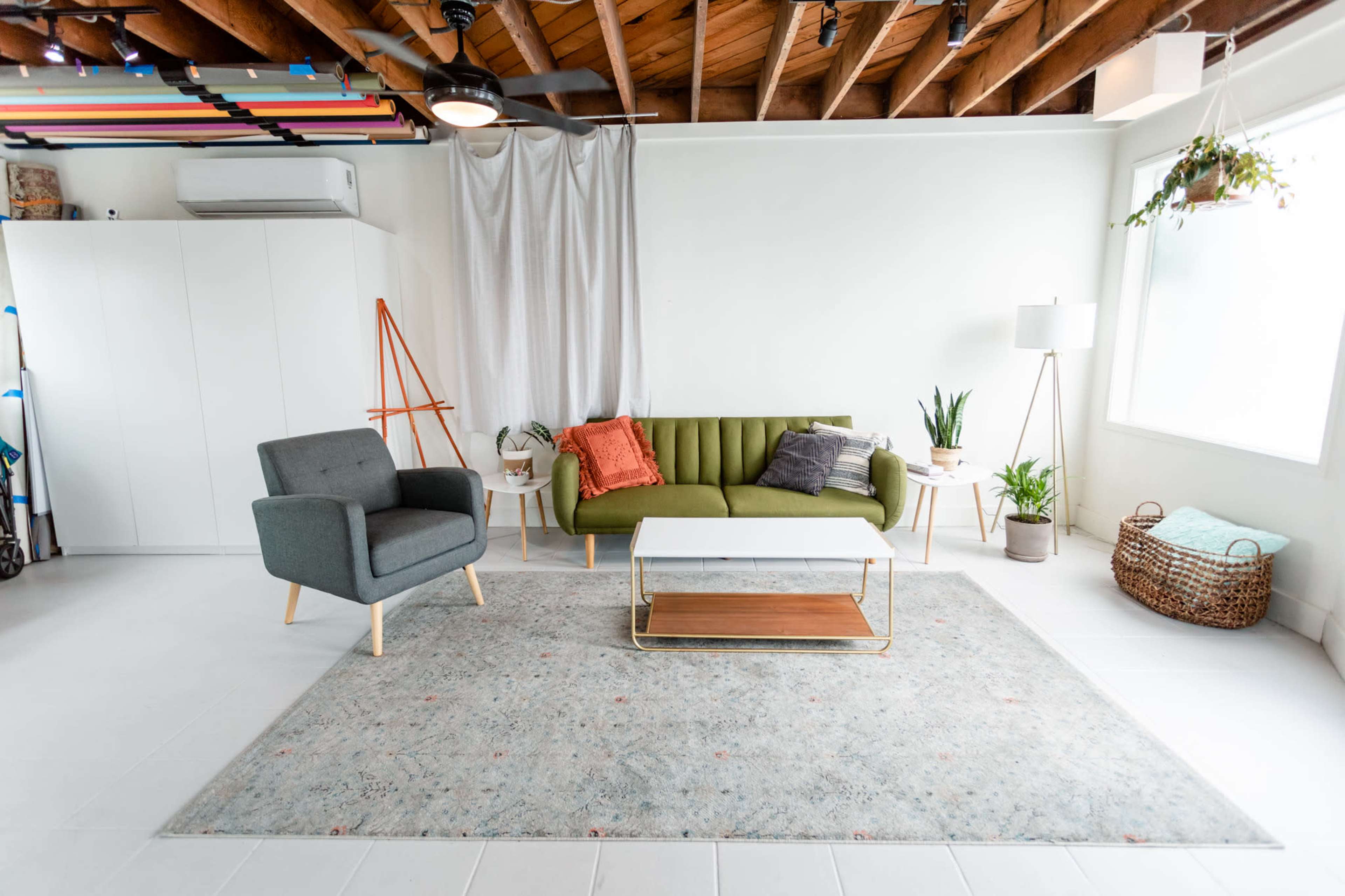 The image shows a minimalist living room featuring a green sofa, a gray armchair, a coffee table, and a large area rug, with plants and natural light from the windows.