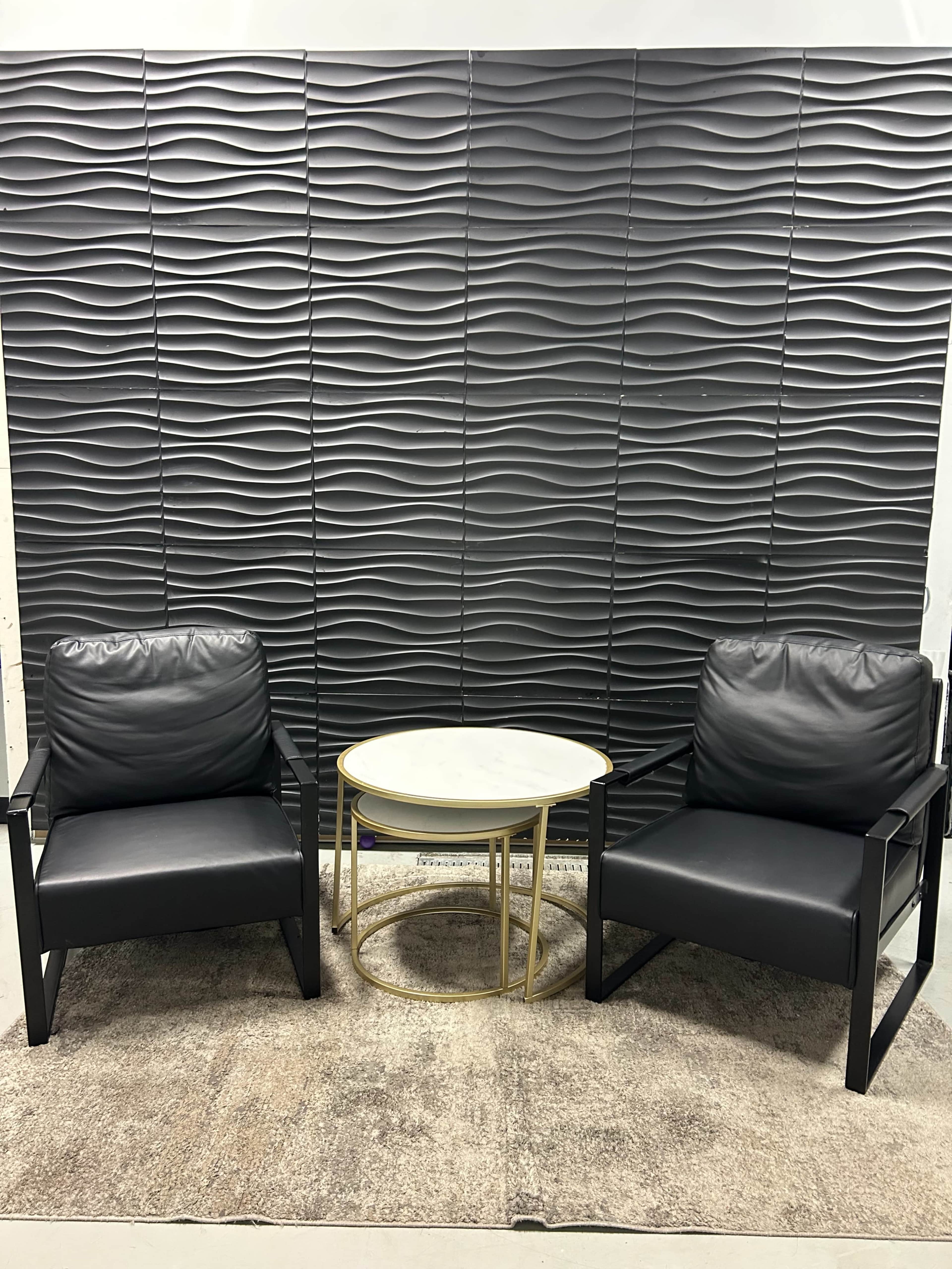 The image shows two black armchairs positioned on a light rug facing a circular gold and white marble table against a textured gray wall.