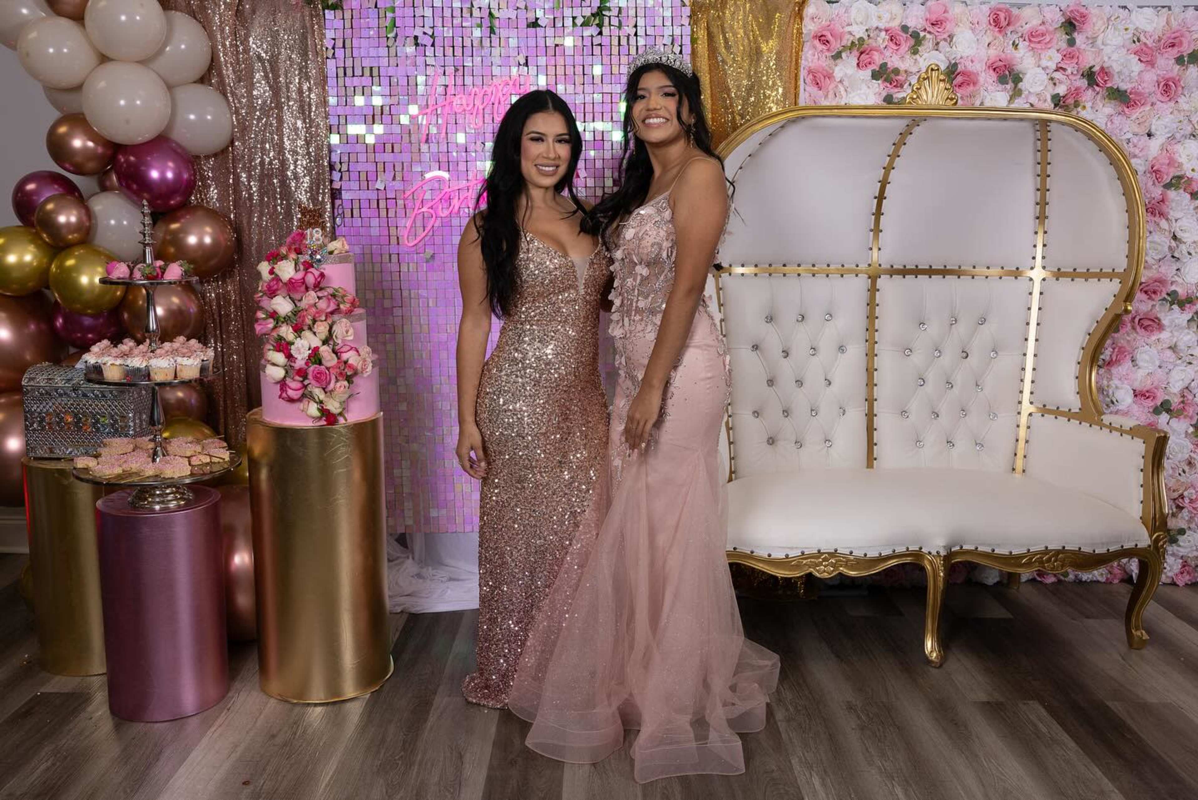 Two women pose in front of a decorative backdrop featuring balloons and floral arrangements, with a shiny cake and a stylishly designed chair nearby.