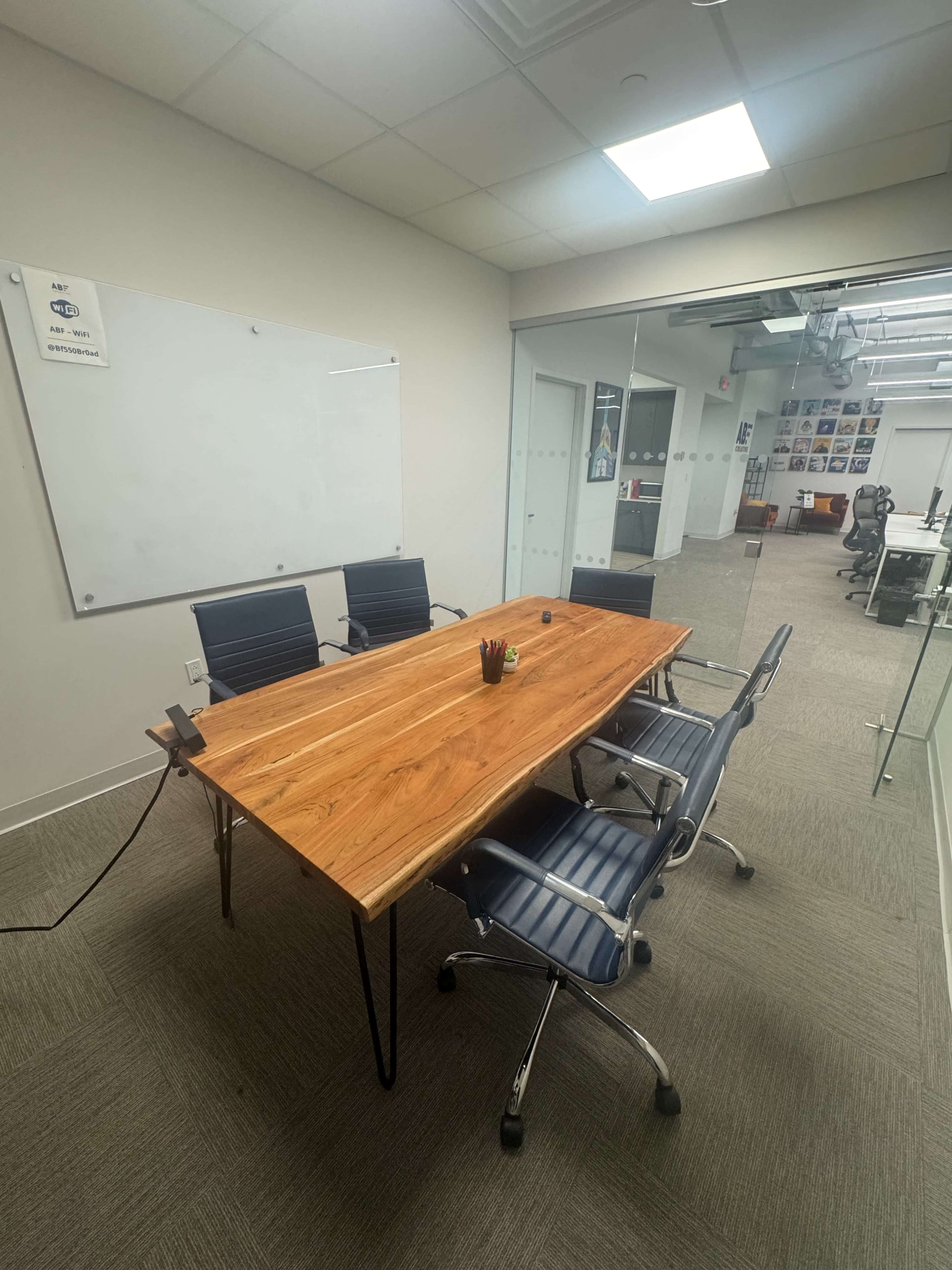 The image shows a conference room with a wooden table and four chairs, featuring a whiteboard on one wall and glass partitions.