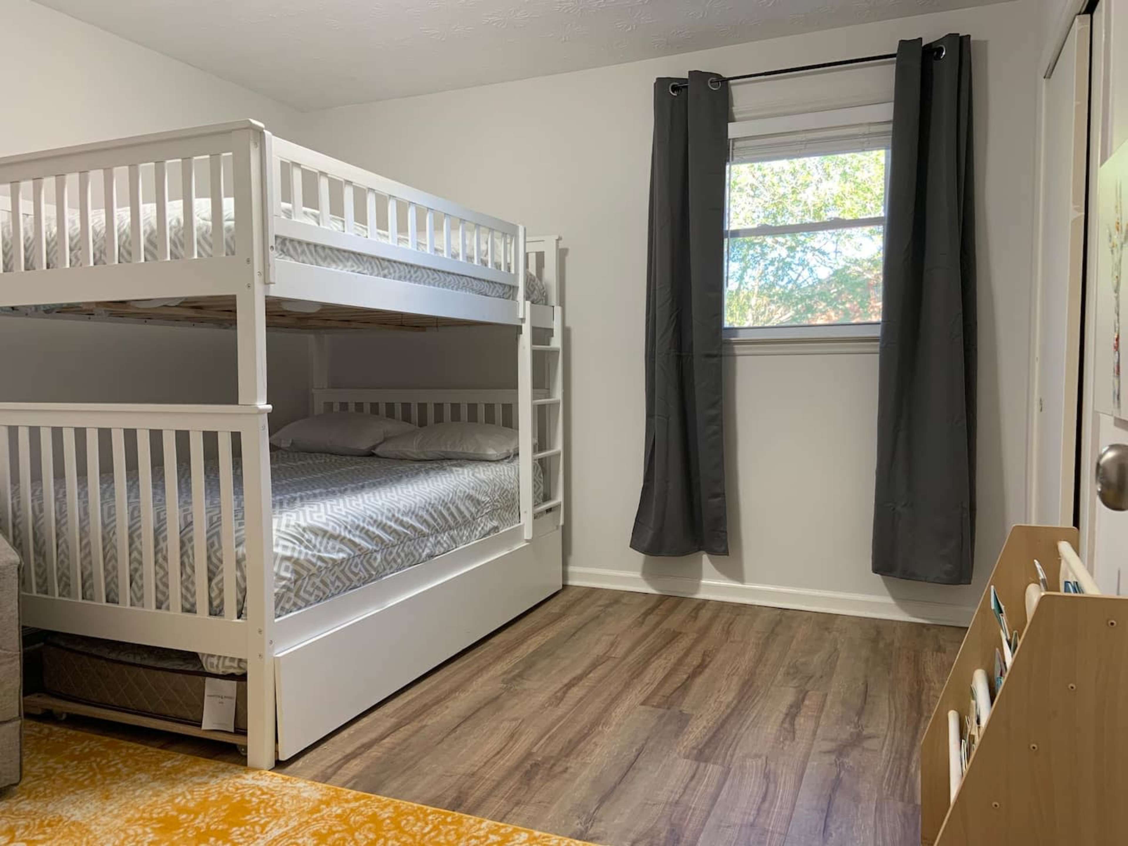 A well-lit room features a bunk bed against a white wall, with a window and gray curtains, along with a small bookshelf and carpeted floor.