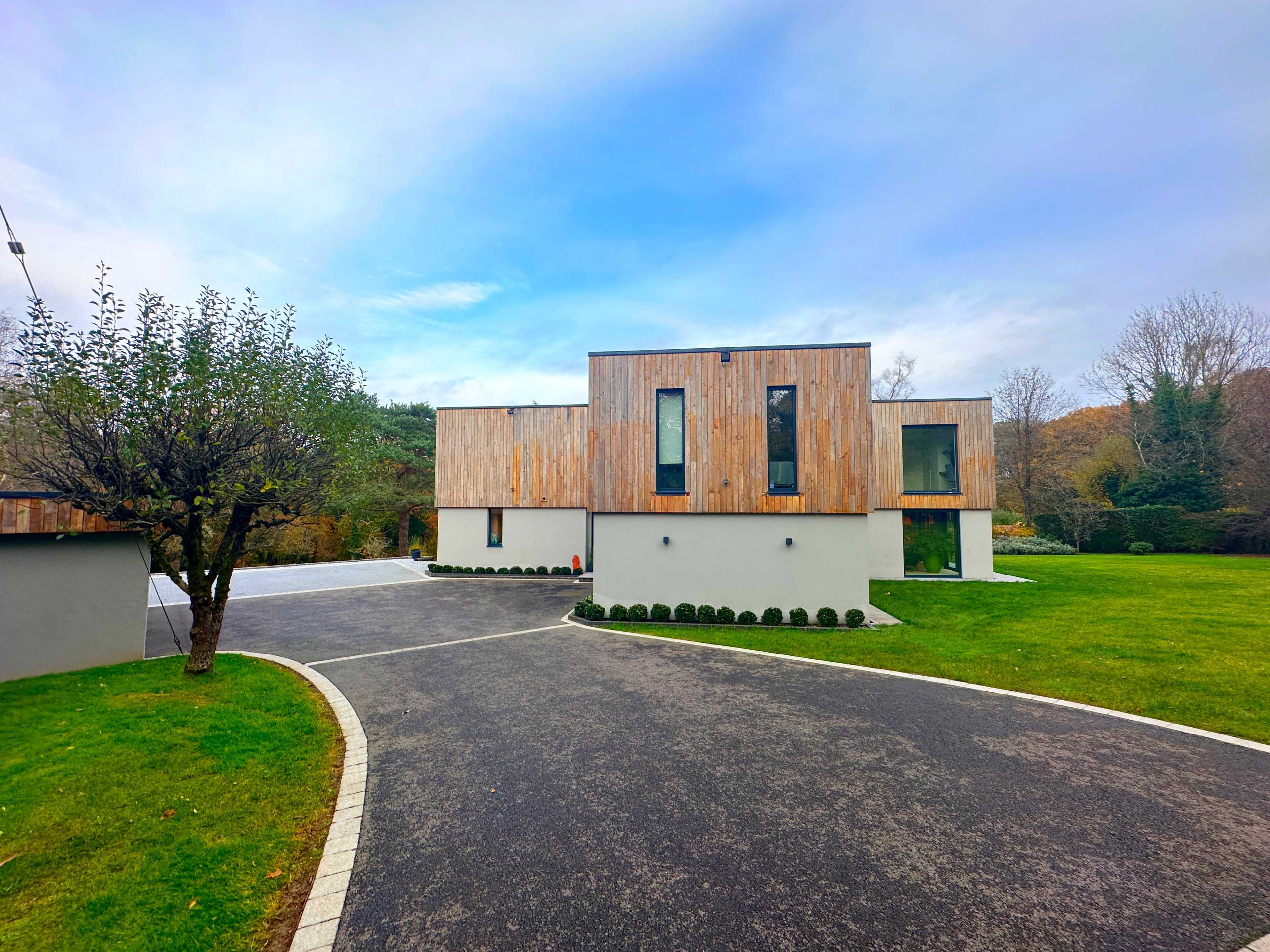 A modern two-story house with a wooden facade sits beside a manicured lawn and a paved driveway.