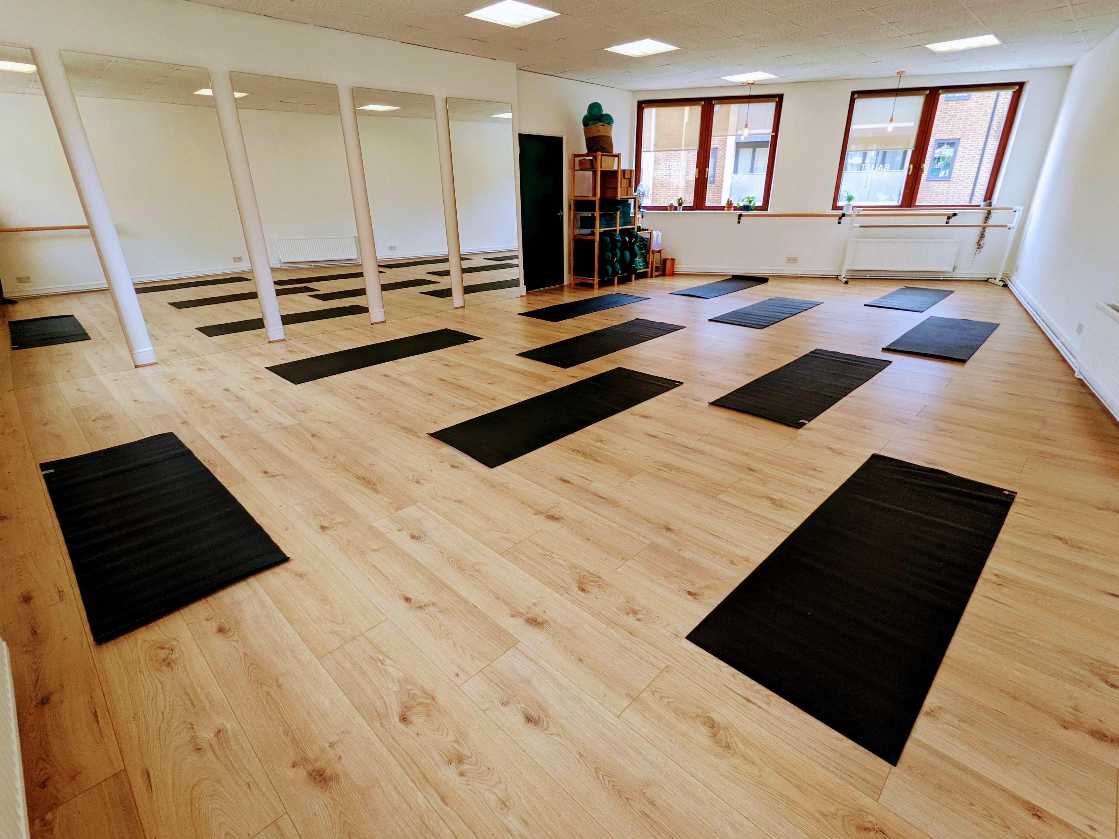 A well-lit yoga studio with black mats arranged on a wooden floor, along with large mirrors and shelves in the background.