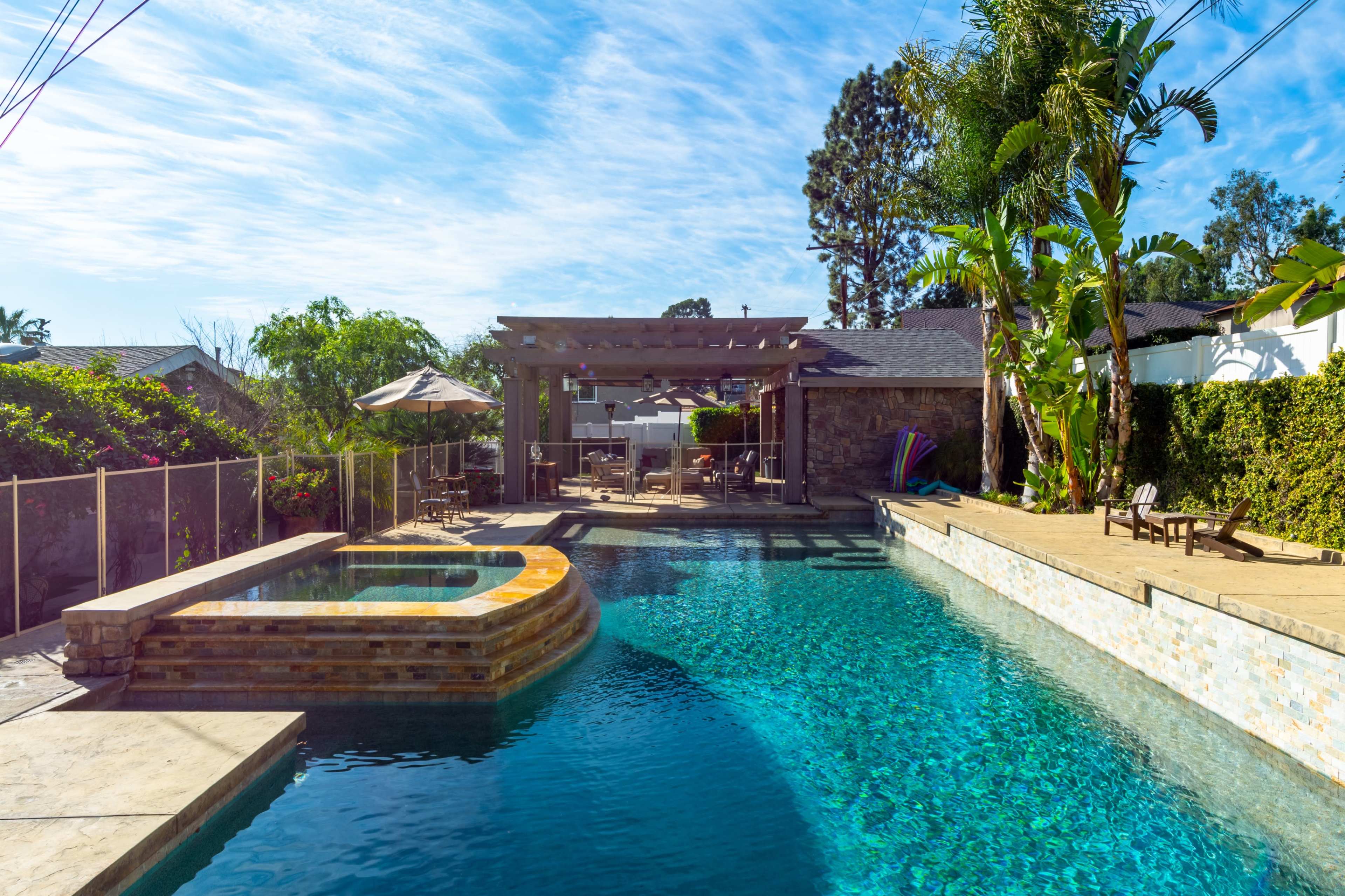 The image shows a rectangular swimming pool with a connected spa, surrounded by landscaped greenery, outdoor seating, and a pergola.
