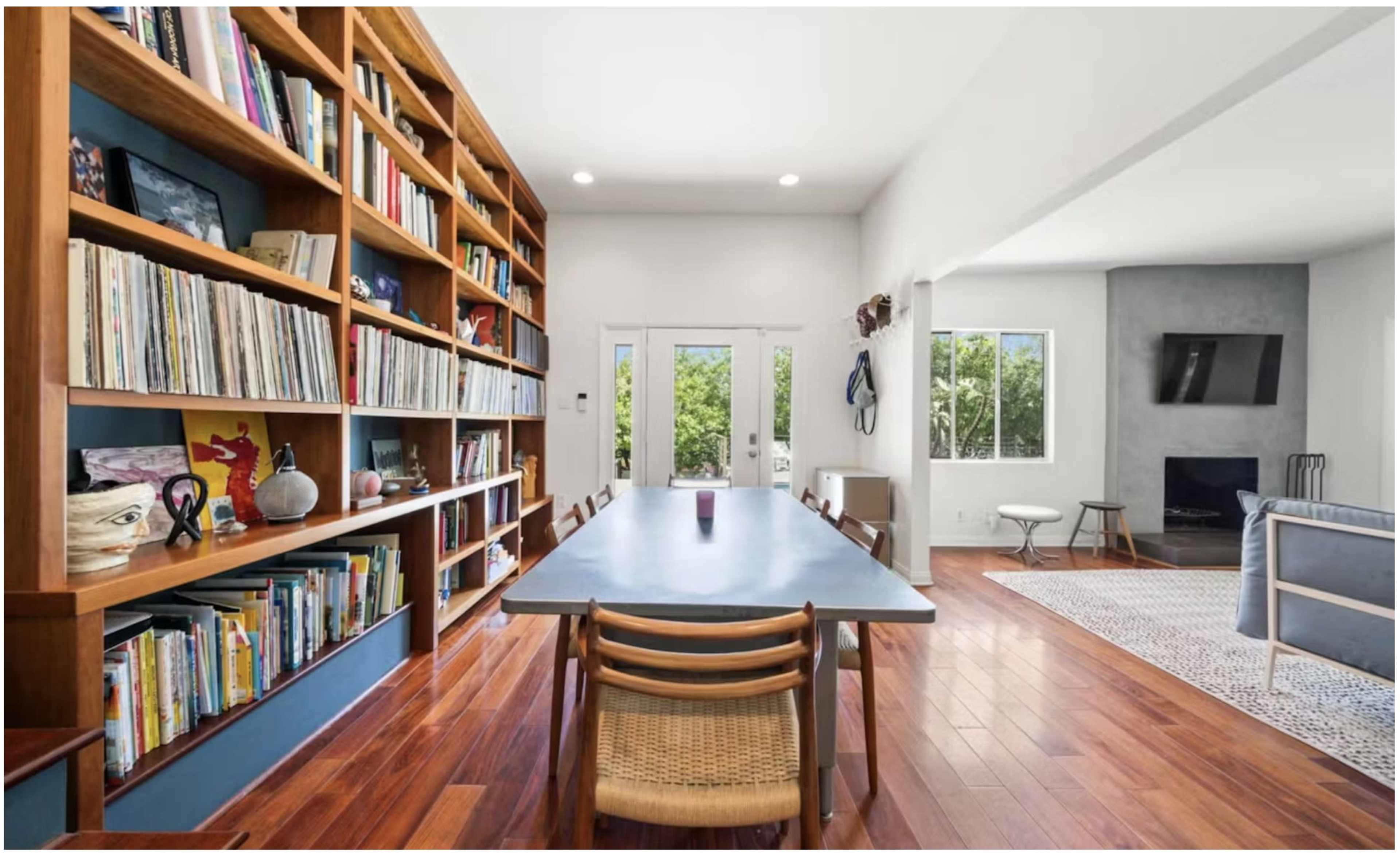 A spacious room features a long table surrounded by wooden chairs, with a large bookshelf filled with records and artwork along one wall, and a fireplace visible at the far end.