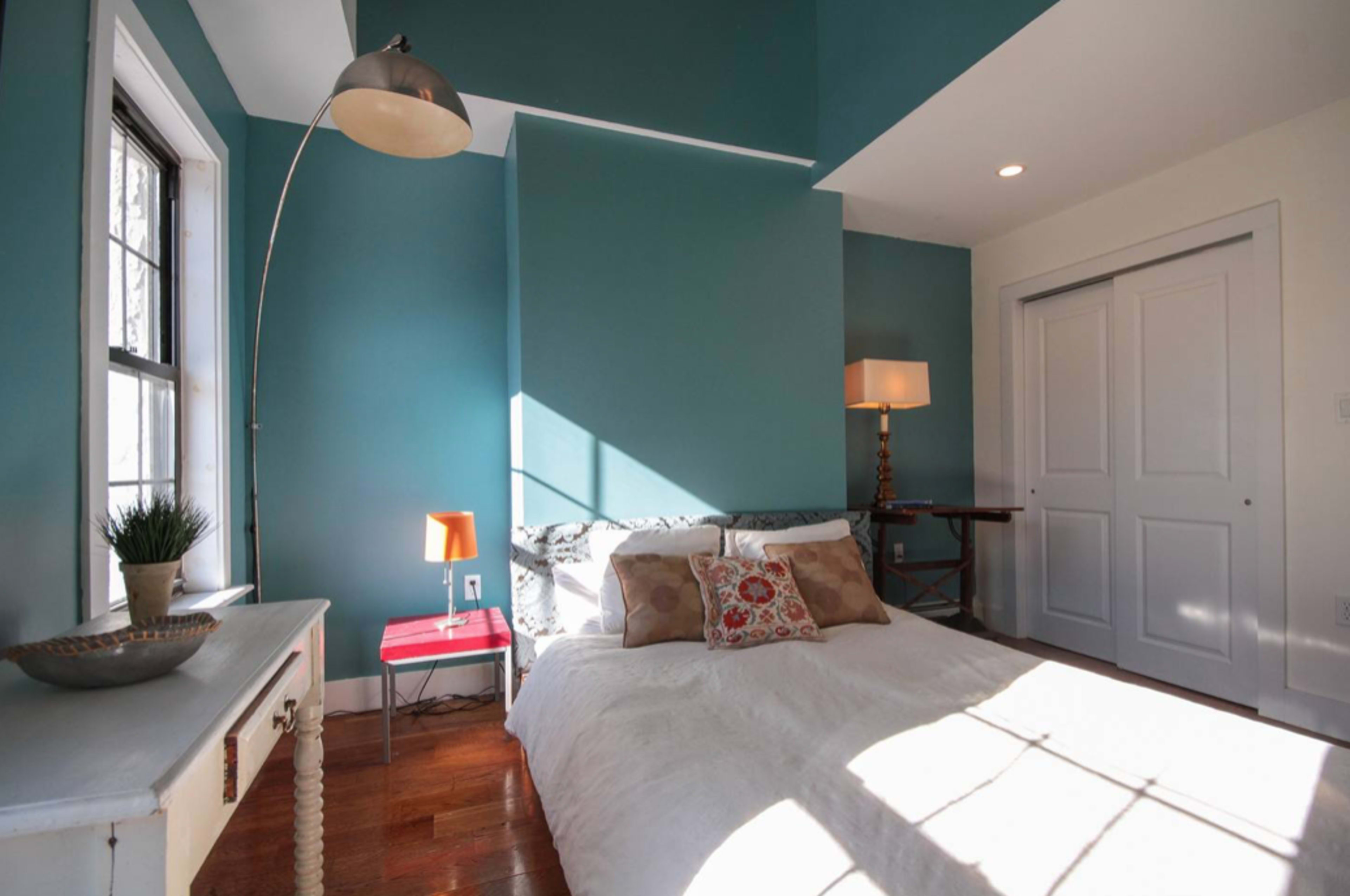 A well-lit bedroom featuring a bed with decorative pillows, a floor lamp, a bedside table with a small lamp, and a closet door.