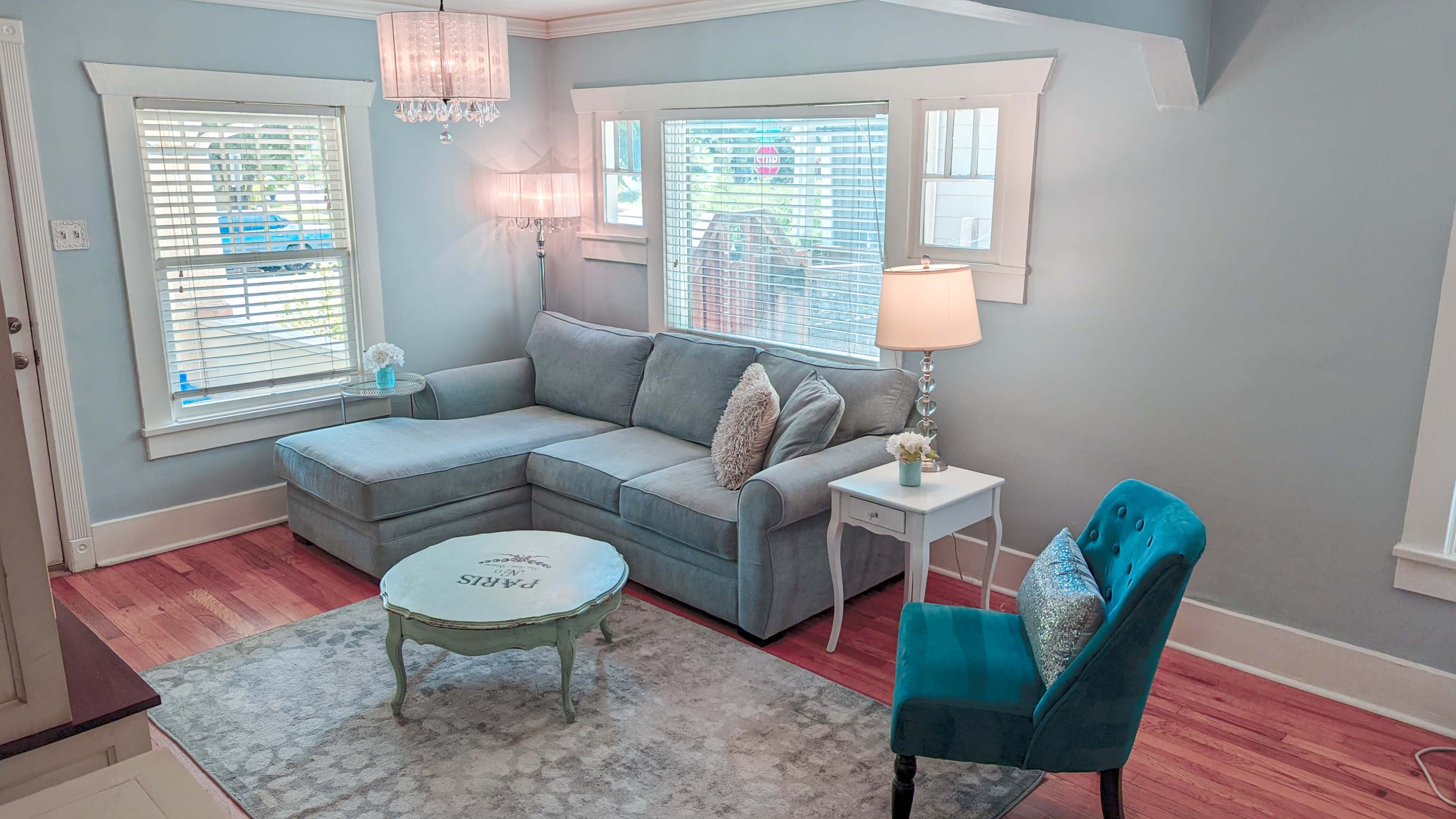 A light blue living room featuring a sectional sofa, a small round coffee table, a lamp, and a decorative armchair, all illuminated by natural light.