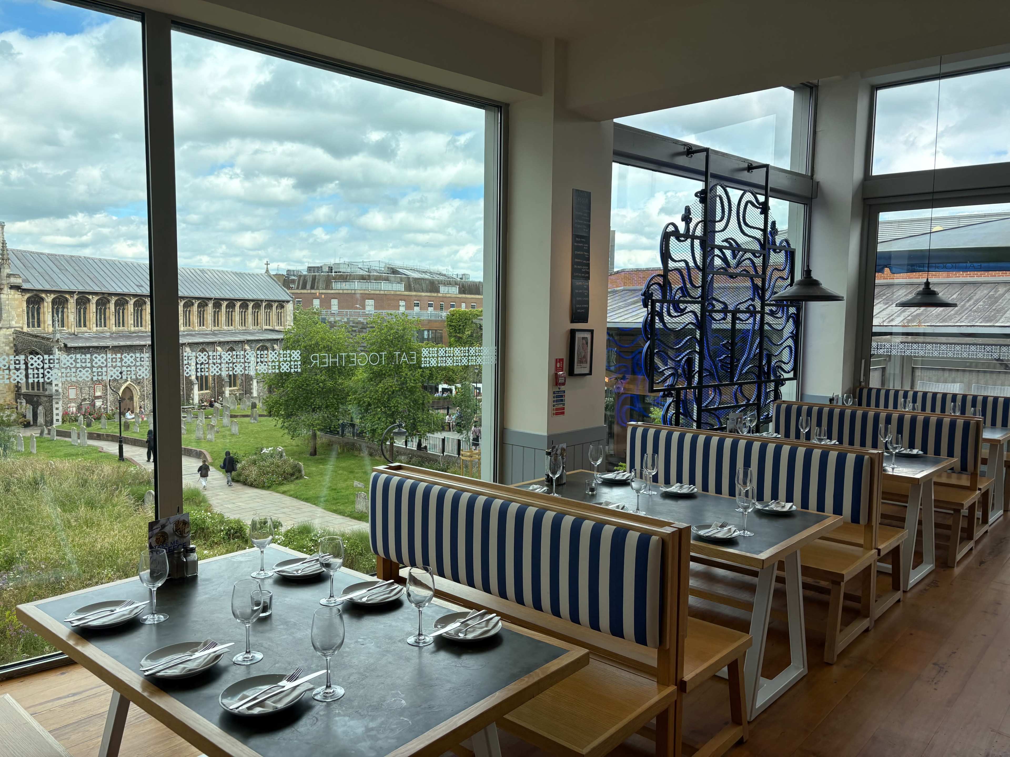 The image shows a modern restaurant interior with striped benches and tables set for dining, overlooking a courtyard and historic building through large windows.