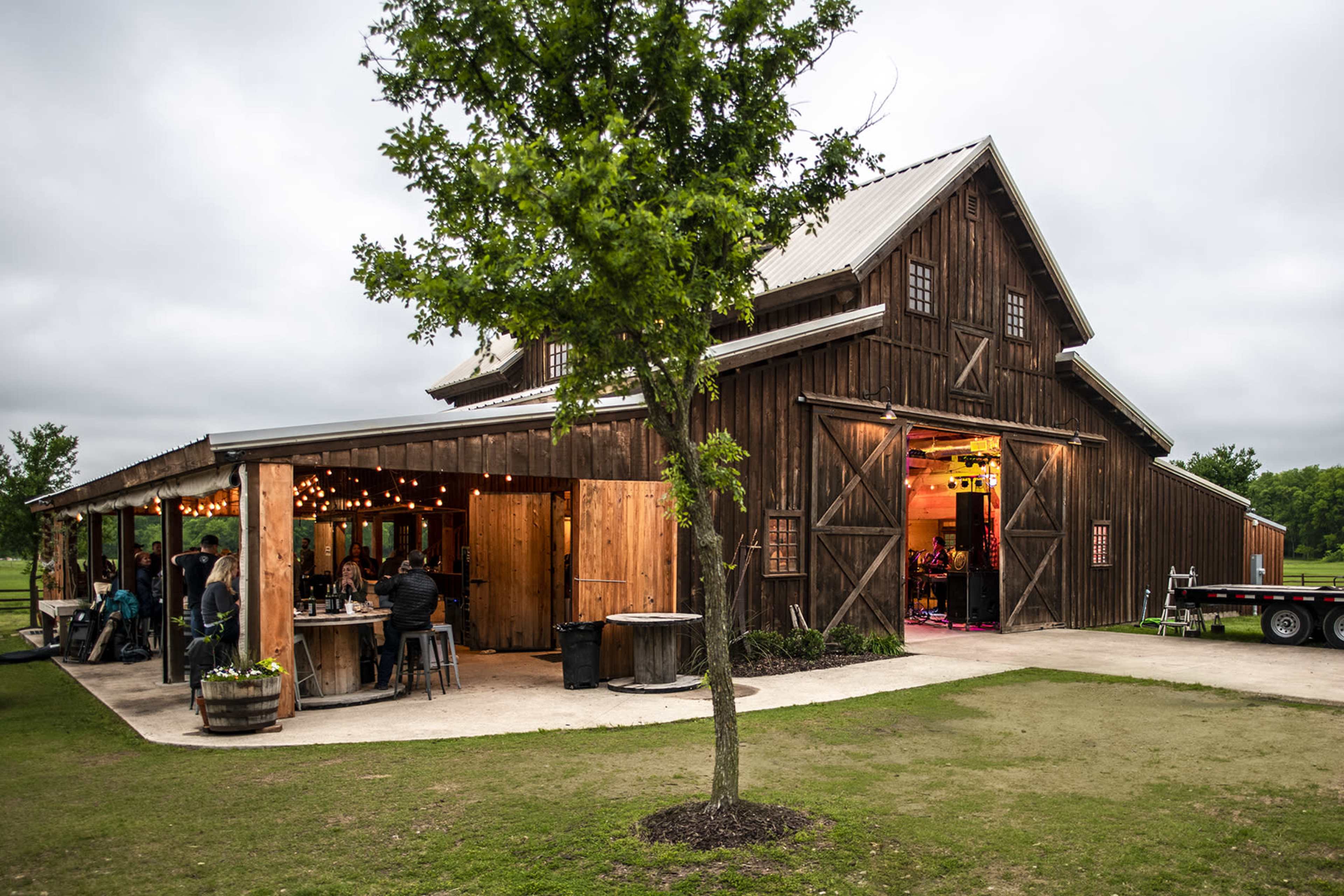 A rustic wooden barn with a covered outdoor area, adorned with string lights and surrounded by a grassy lawn.