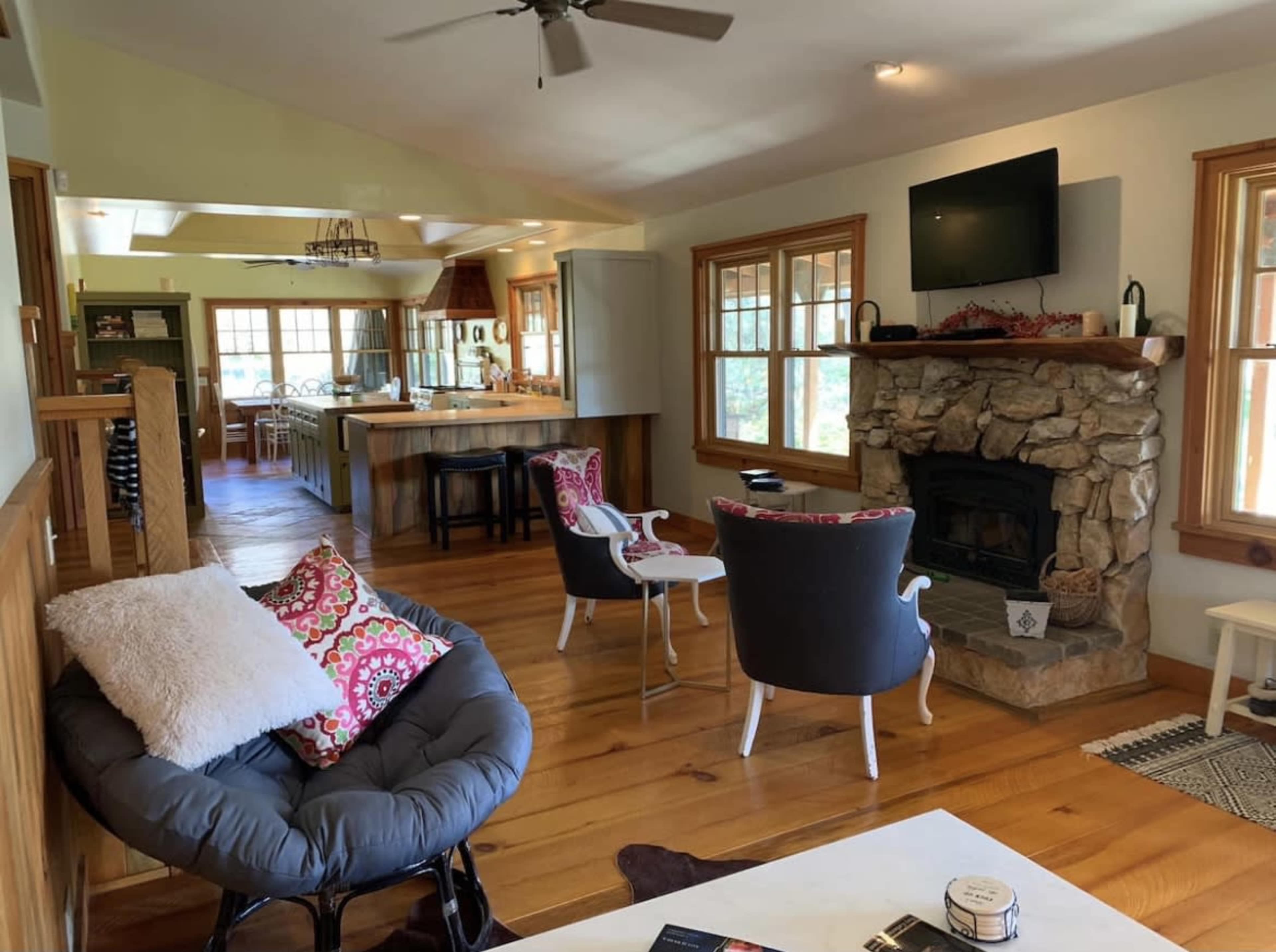 A spacious living area with wooden flooring, a stone fireplace, and comfortable seating arranged around a coffee table, leading to a kitchen in the background.