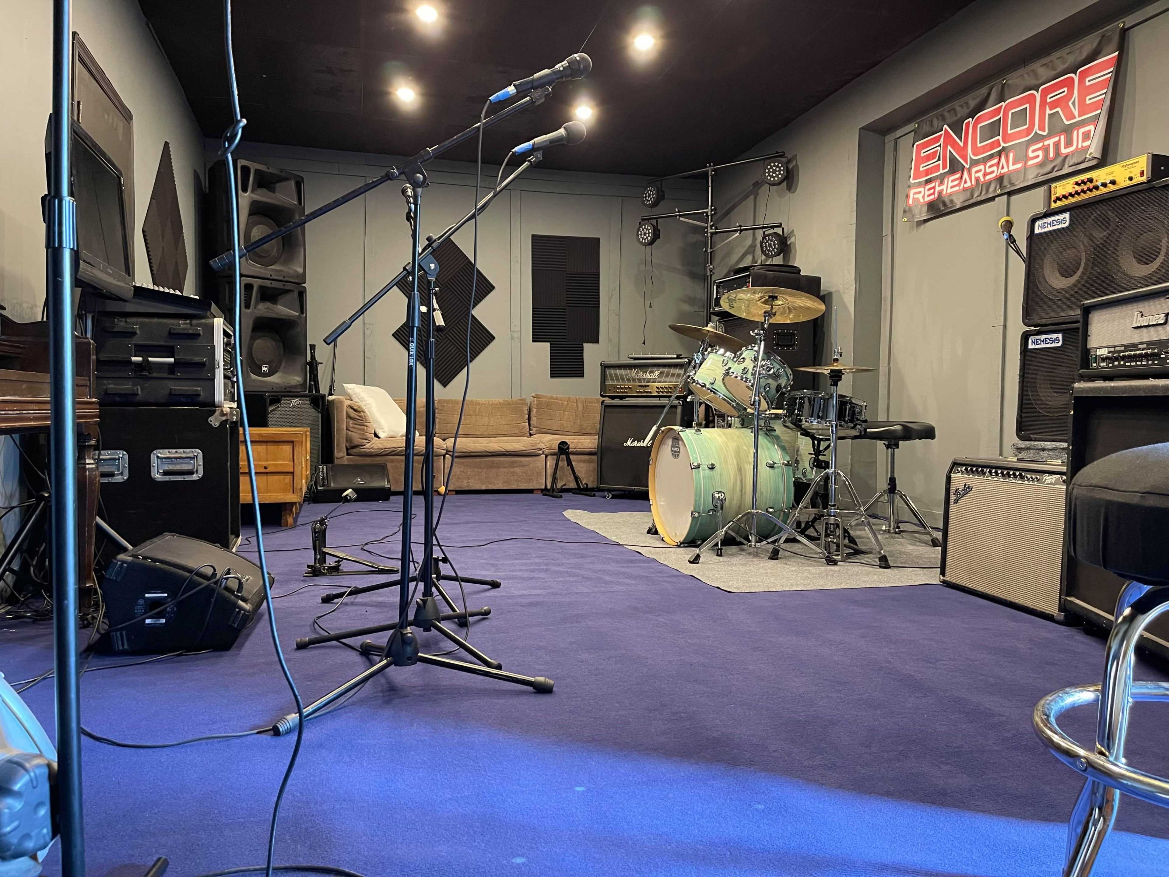 A music rehearsal studio with microphones set up, a drum kit, amplifiers, and a sofa in a soundproofed room.