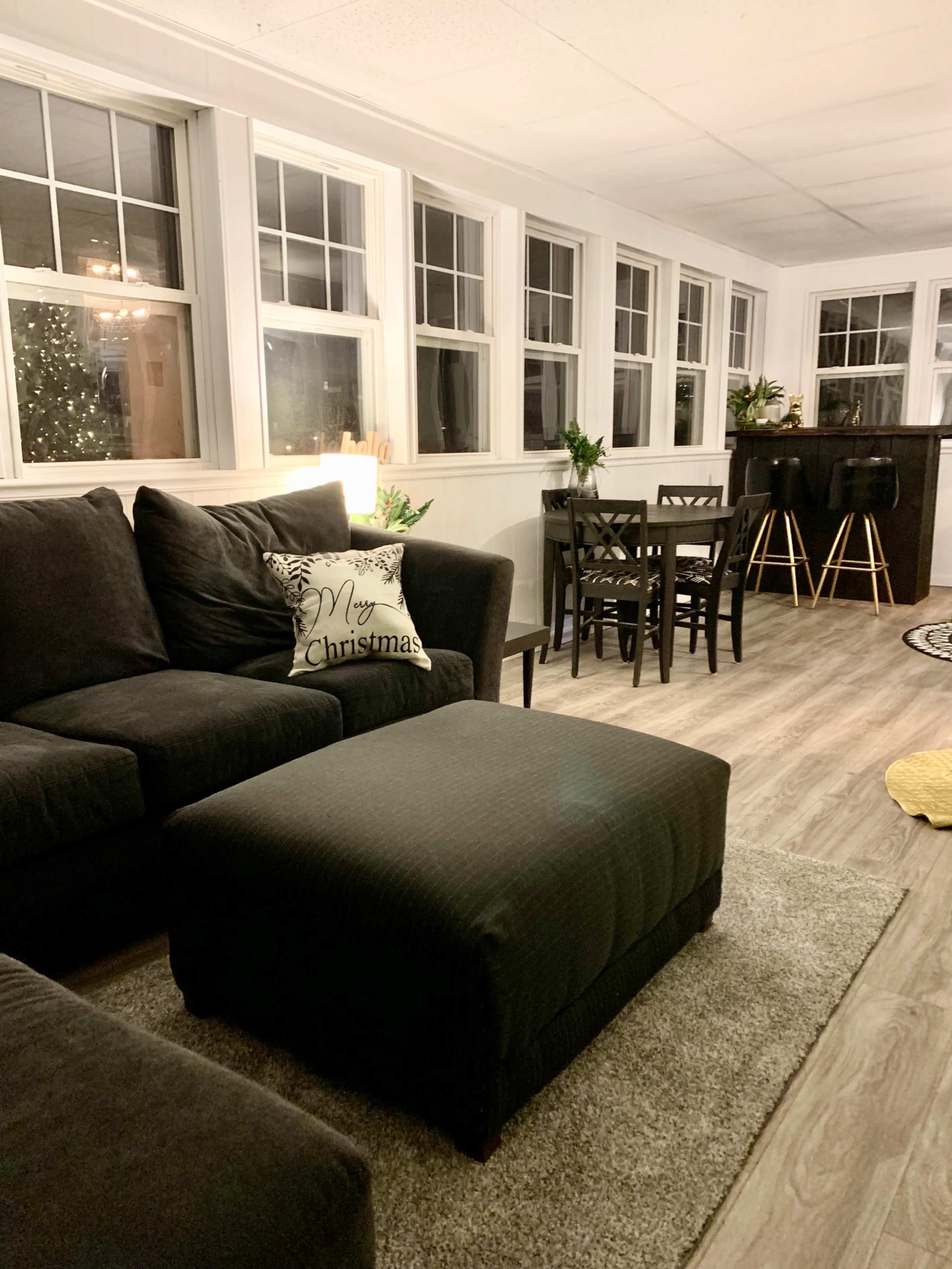 A cozy sunroom with a dark couch, a decorative pillow reading "Merry Christmas," and a dining area with black chairs and a tall table.