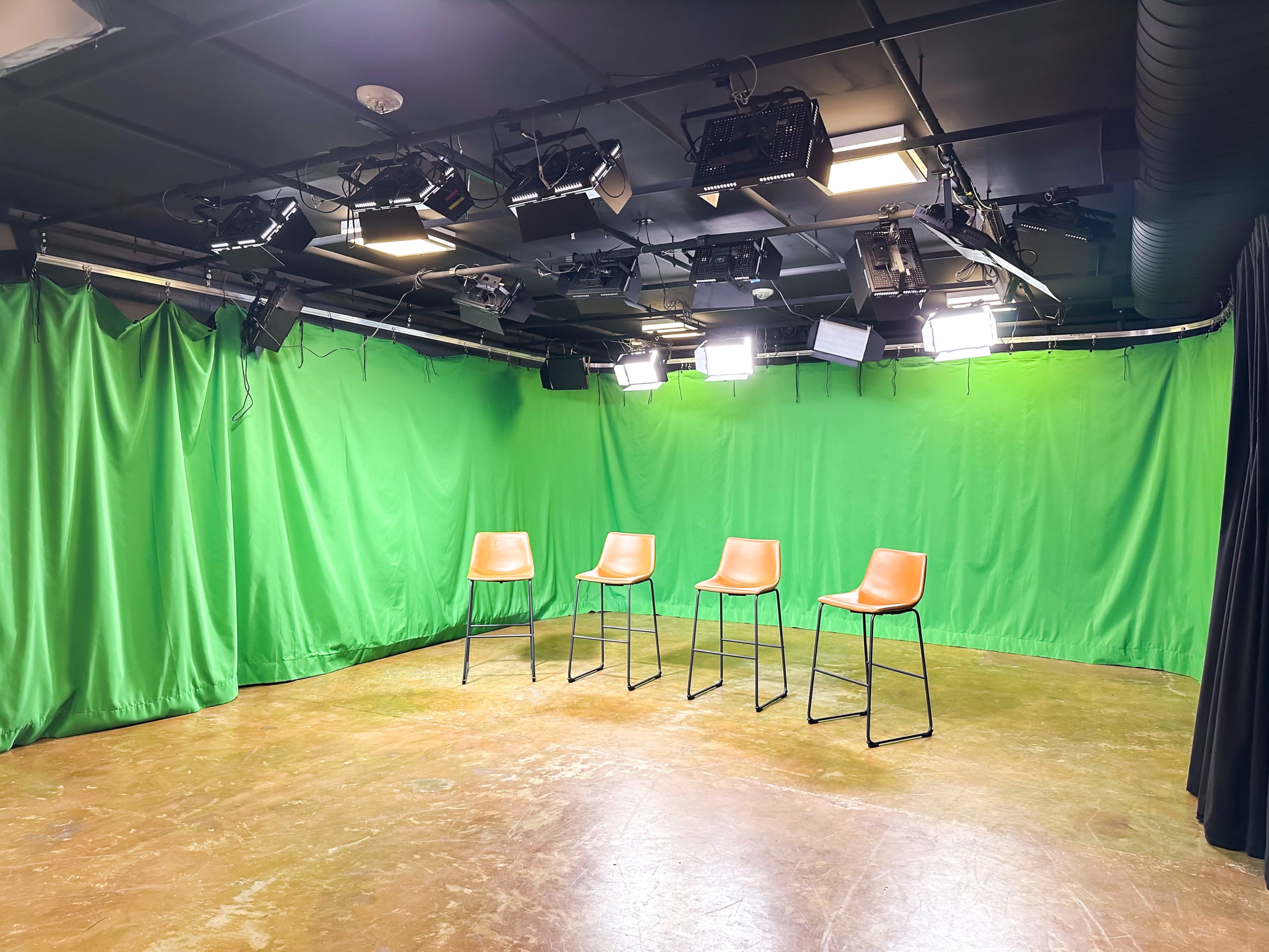 A filming studio with four orange chairs arranged in front of a green backdrop and several overhead lights.