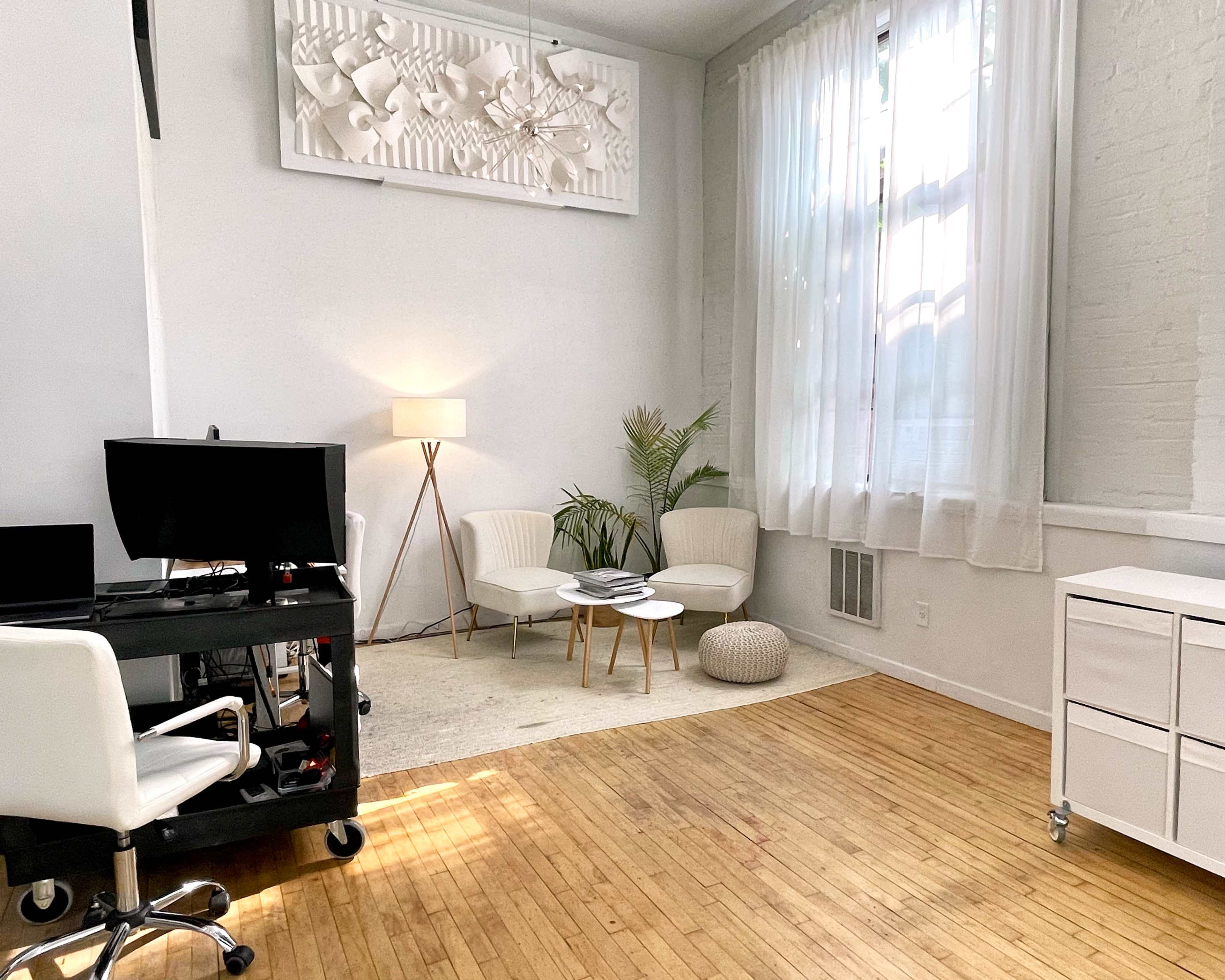 A bright, minimalist office space featuring two modern chairs, a small table, a decorative plant, and a desk with a computer setup.