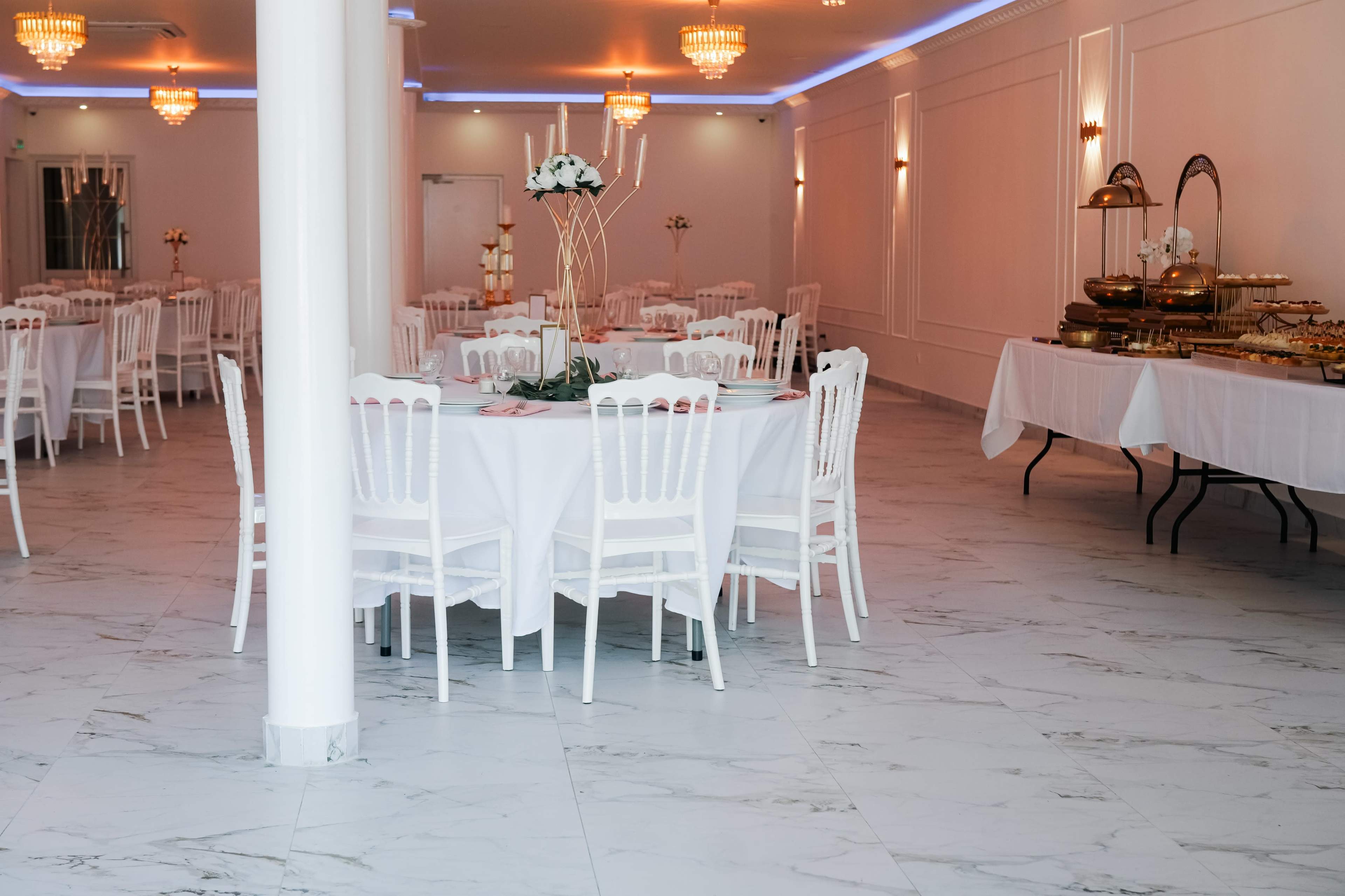 The image shows a spacious dining area with white tables and chairs arranged neatly, marble flooring, and elegant chandeliers overhead.