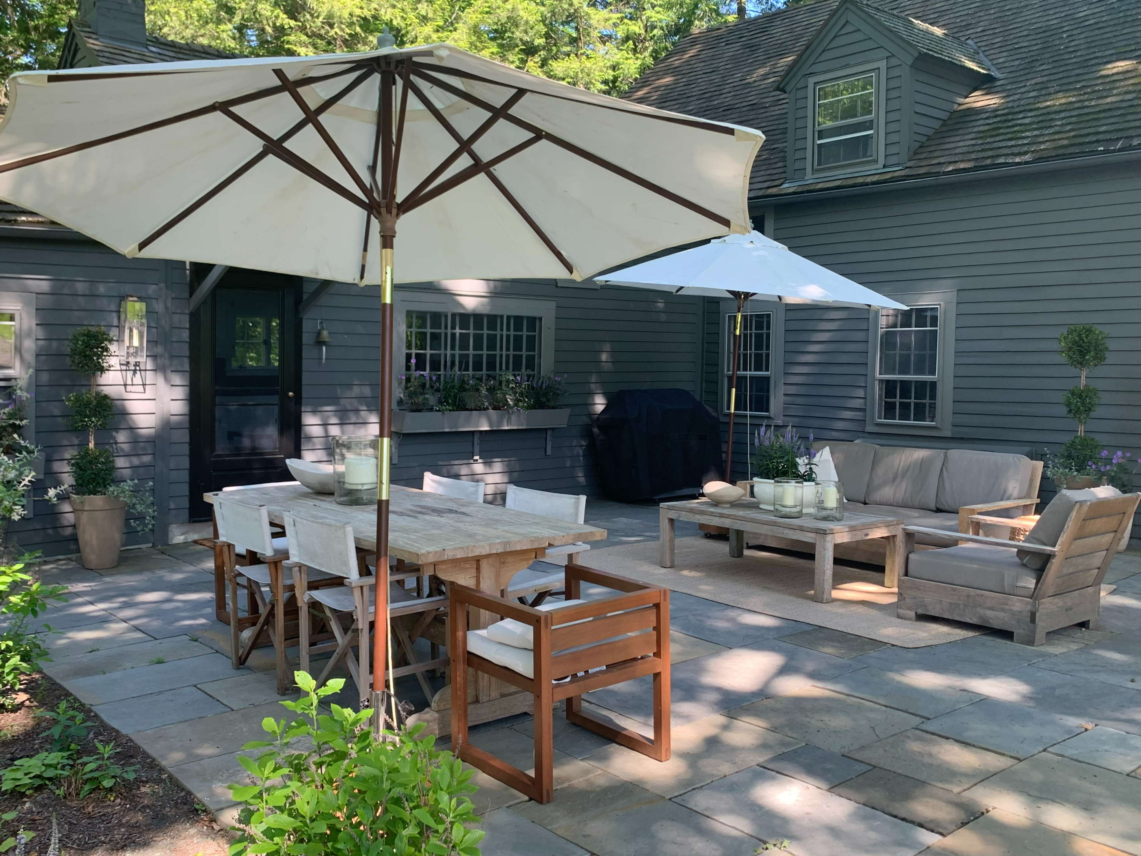 The image shows a stone patio area with a wooden dining table and chairs, two large umbrellas, and a lounge area featuring a sofa and coffee table beside a house with gray siding.