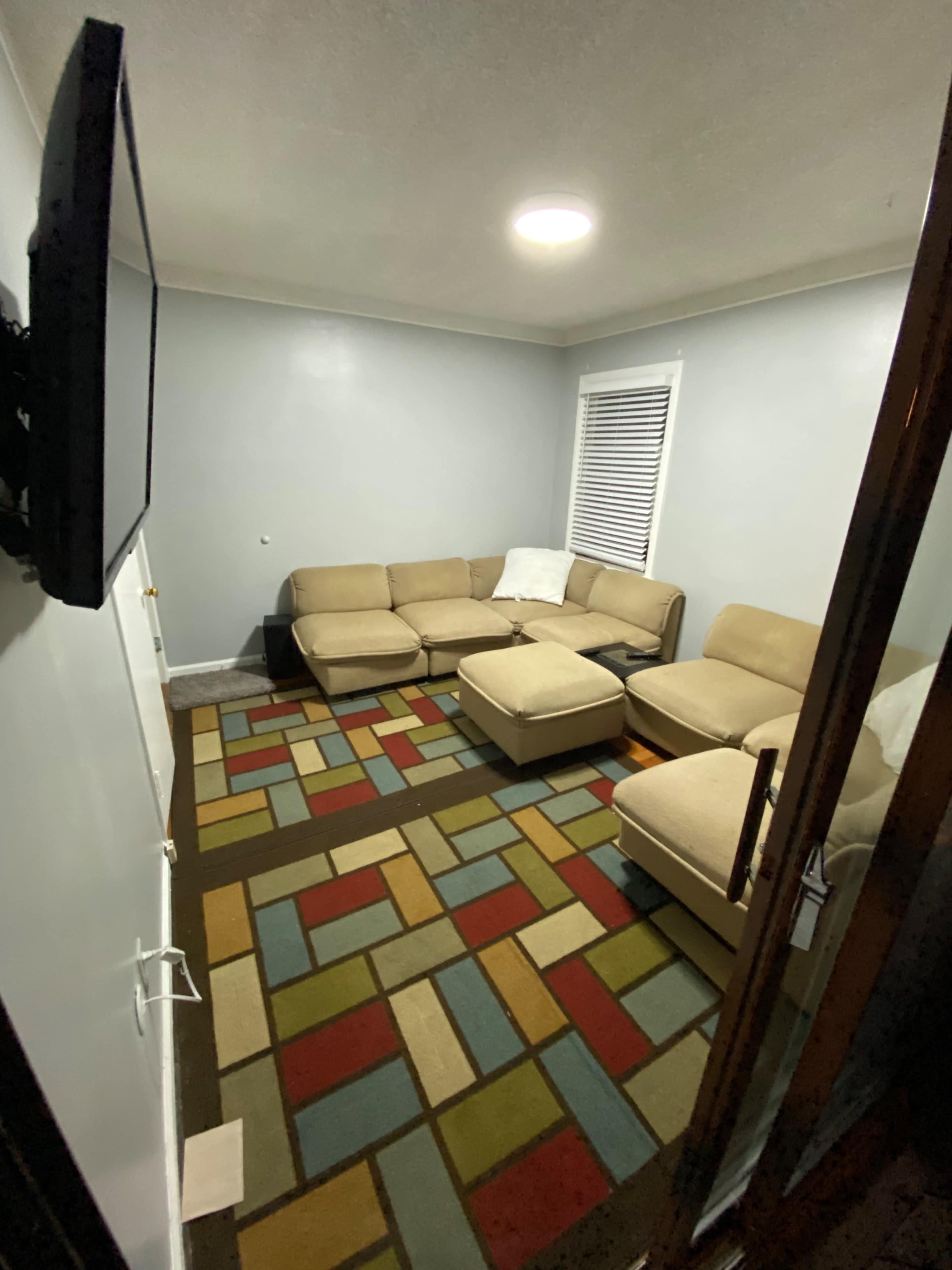 A small living room with a beige sectional sofa, an ottoman, and a multi-colored patterned rug on the floor.