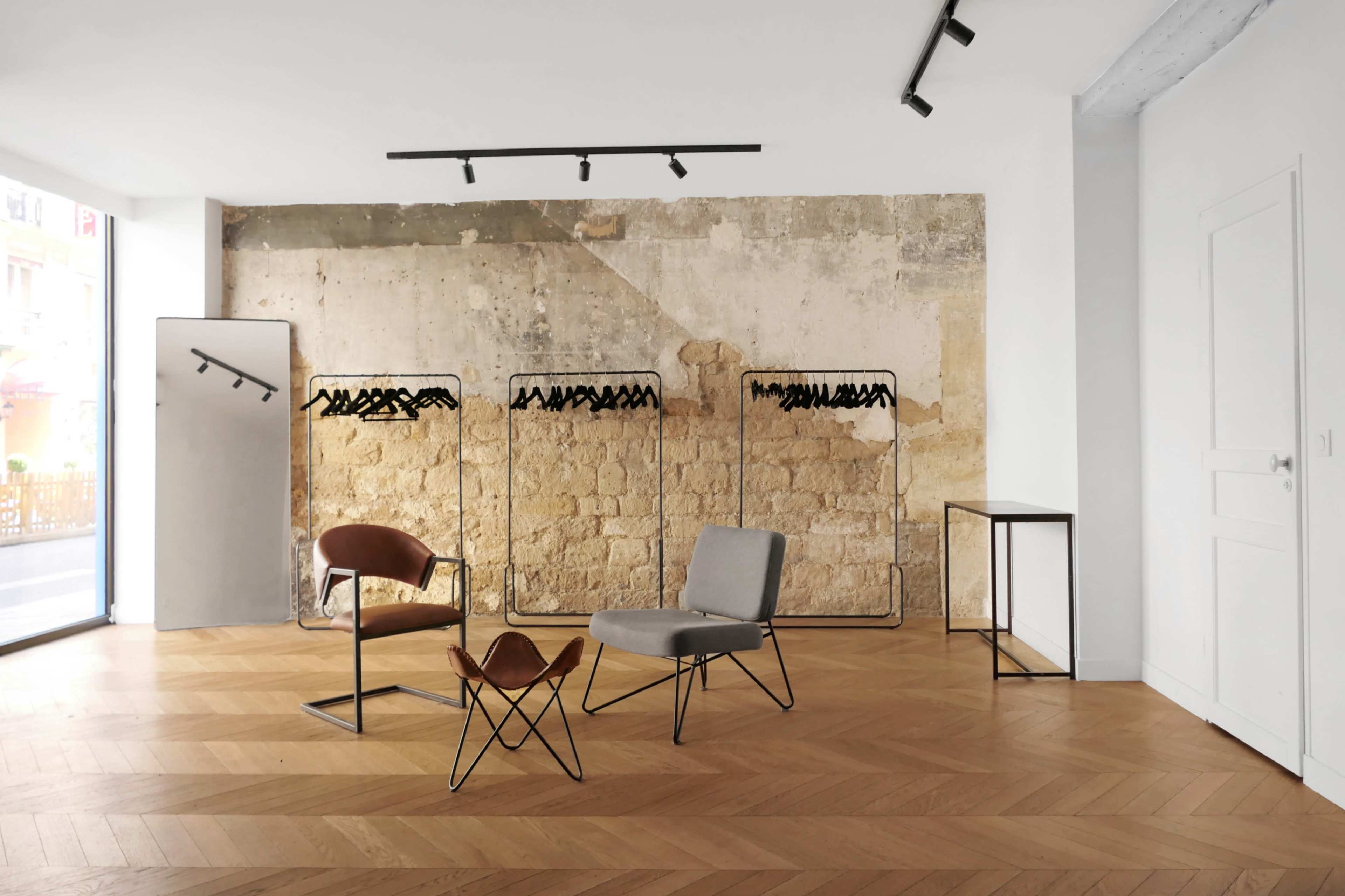 A minimalistic clothing store with two chairs, hanging clothing racks, and a light wooden floor against a textured wall.