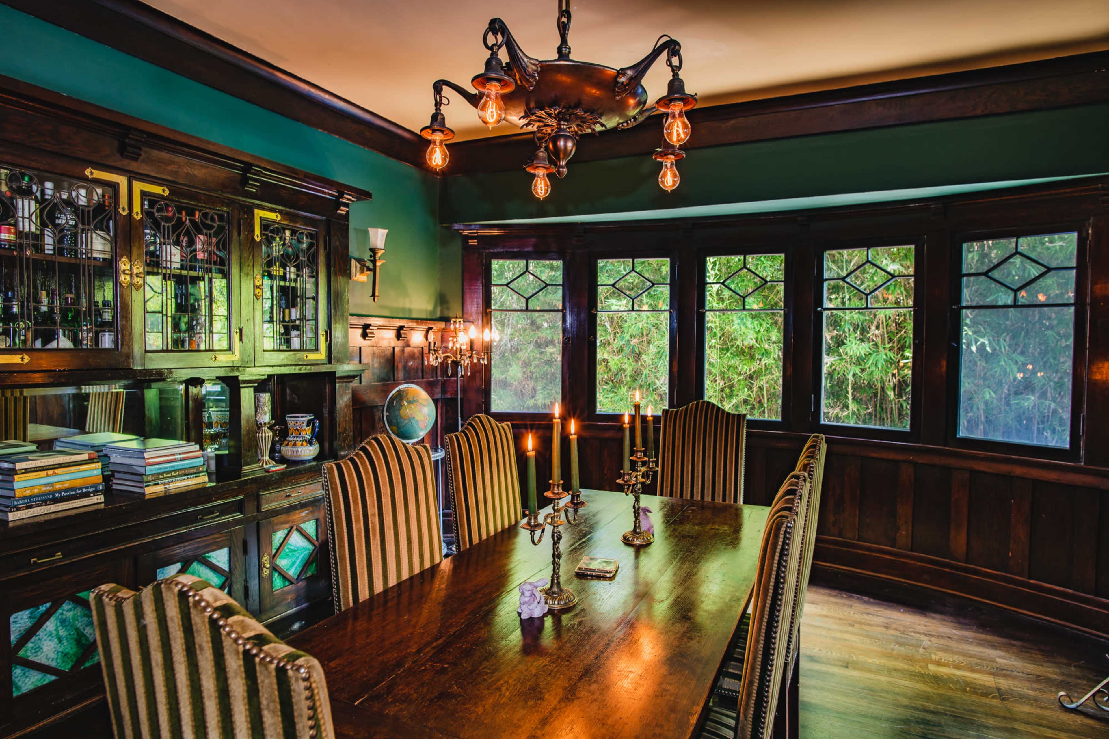A well-furnished dining room with a wooden table, striped chairs, a chandelier, and large windows overlooking greenery.