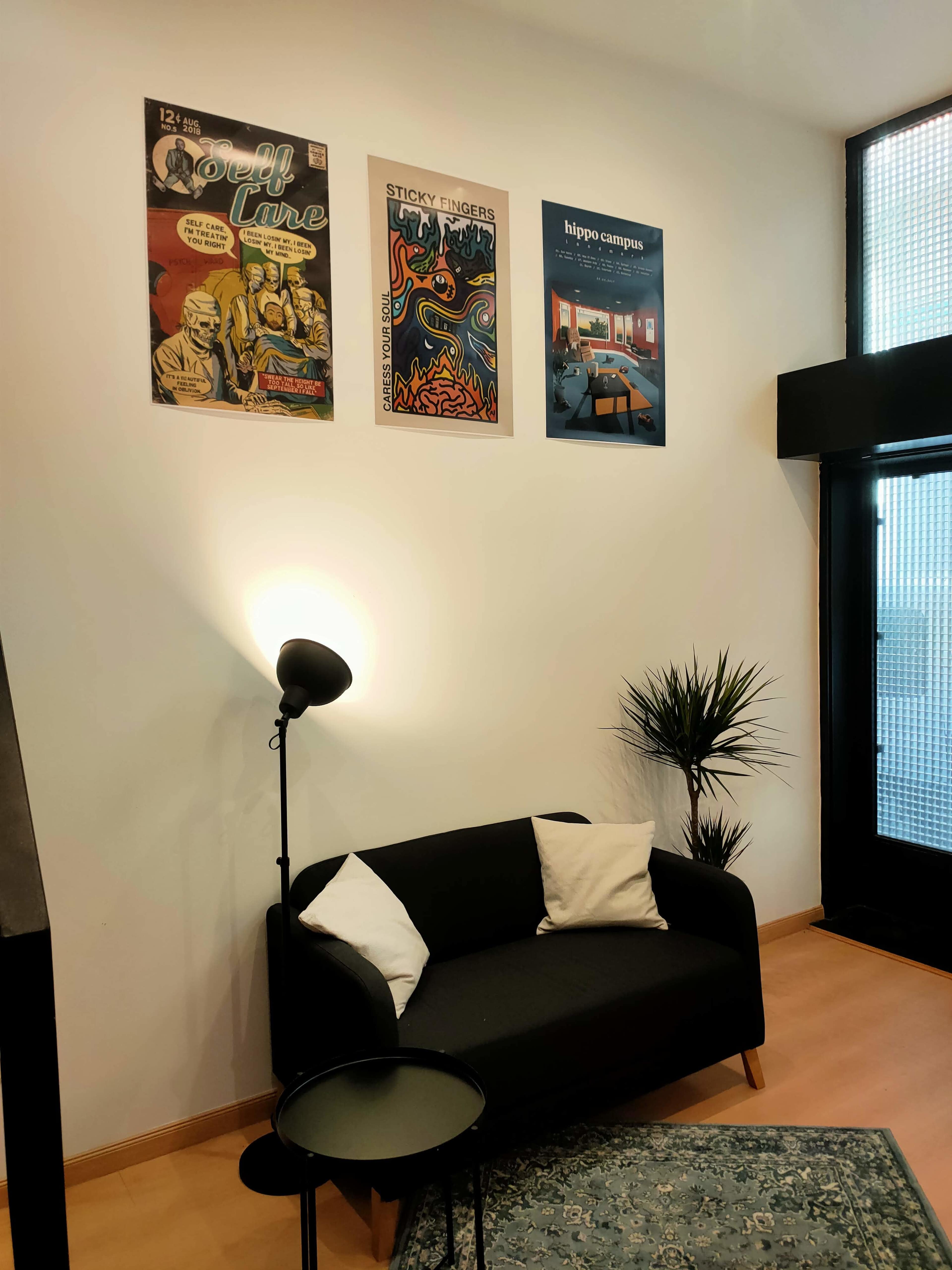 A modern living area with a black couch, a sleek floor lamp, a small round table, and three posters on the wall, along with a potted plant.