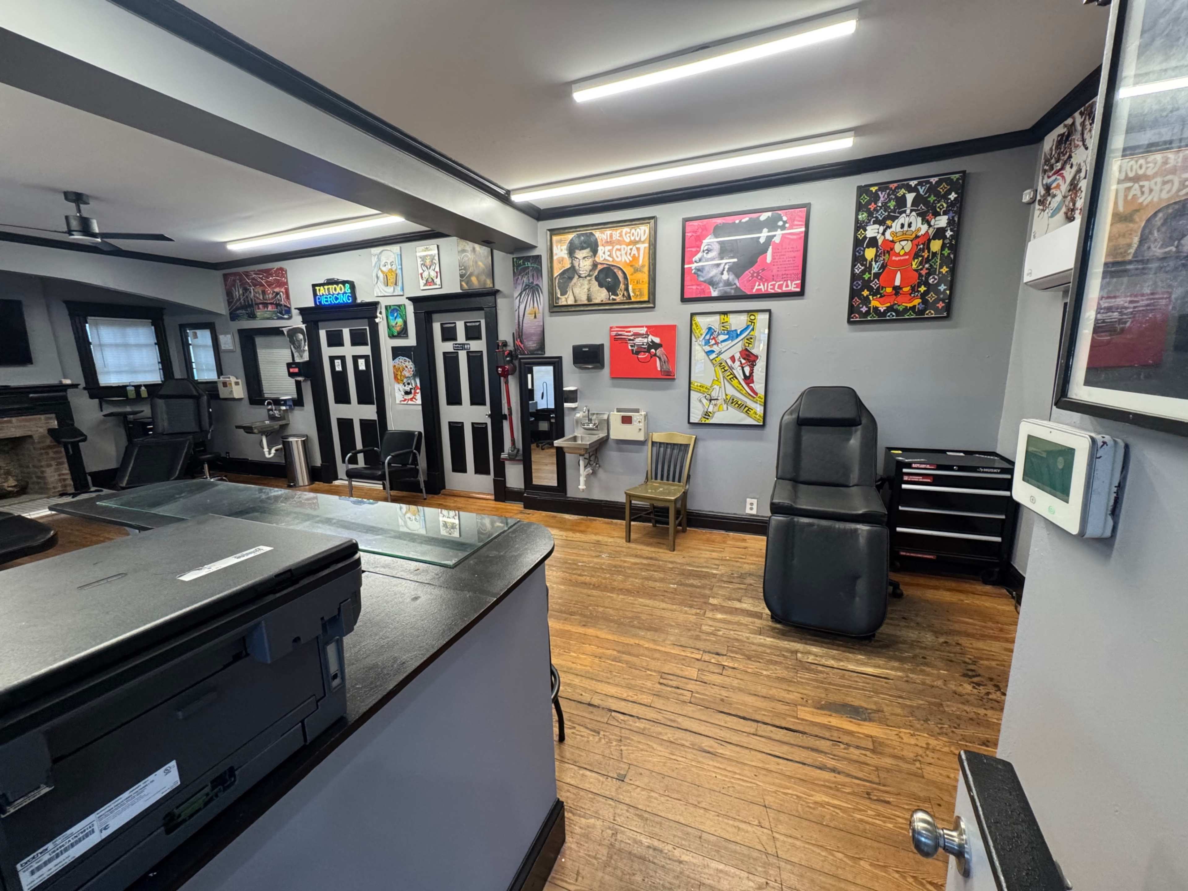 The image shows a modern office or reception area with wooden flooring, various framed artworks on the walls, and an arrangement of office furniture including a chair and printer station.
