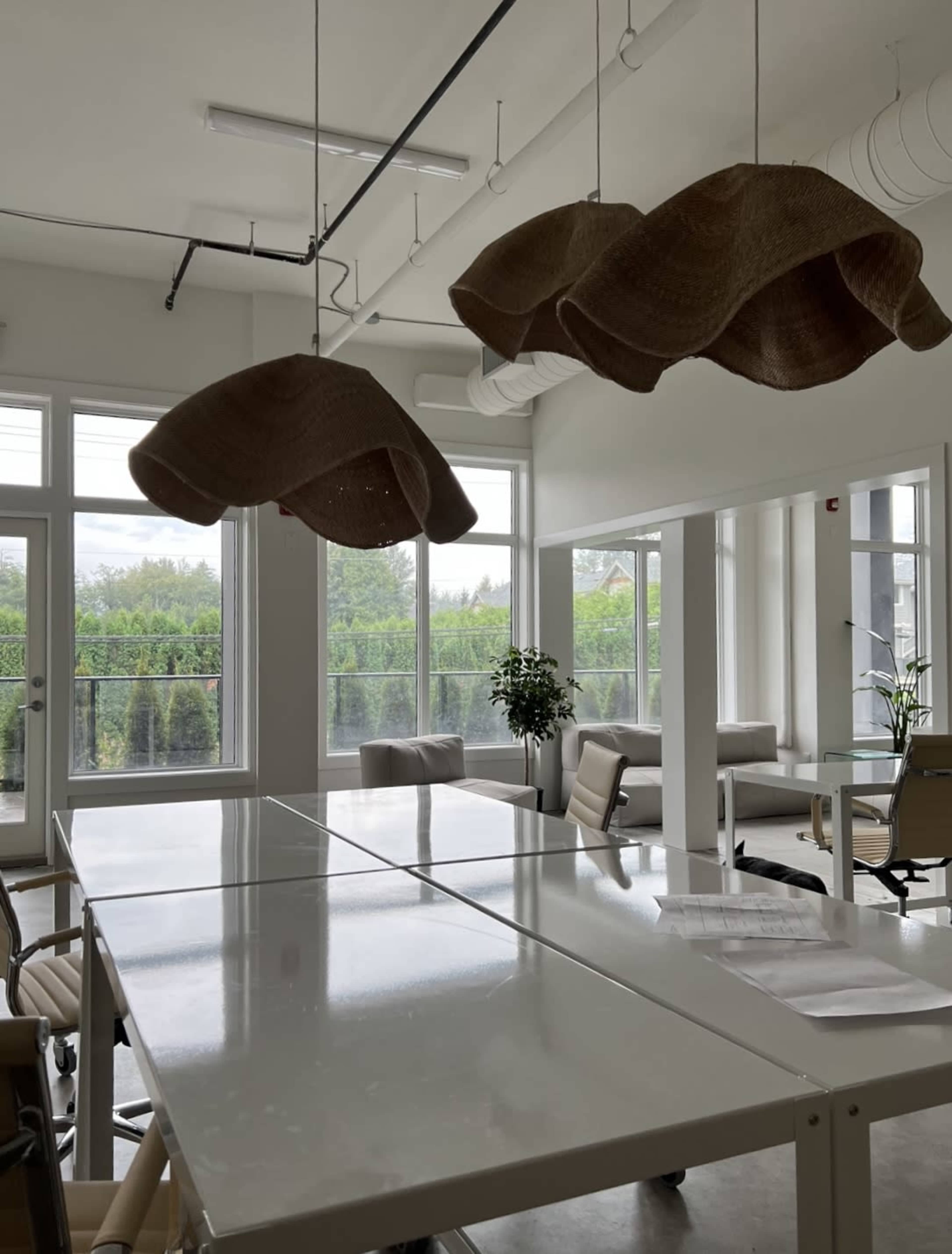 A modern office space with a long white table, two large pendant lights hanging above, and windows overlooking greenery.