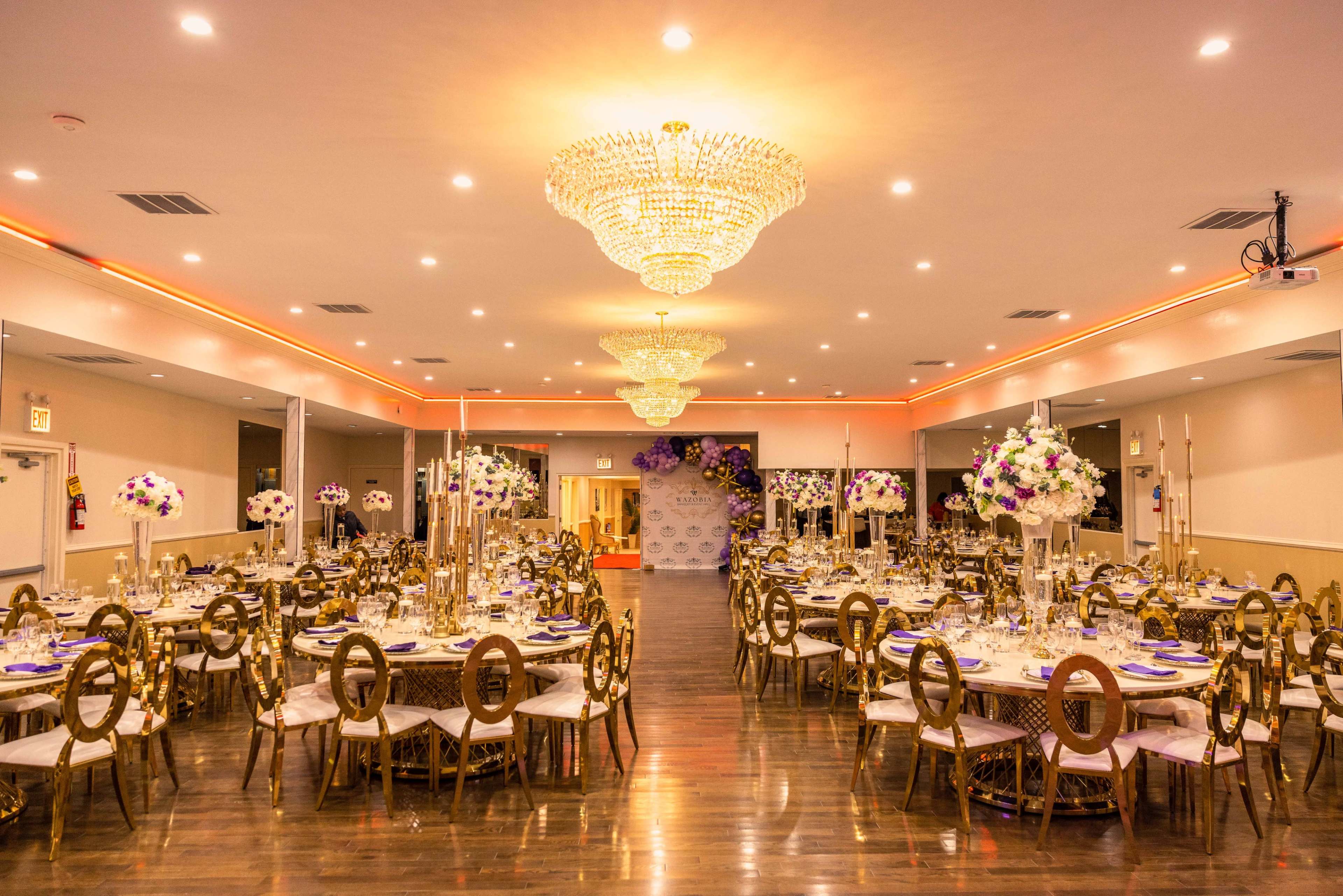 A spacious banquet hall features elegantly set tables with gold chairs, large floral centerpieces, and chandeliers illuminating the space.