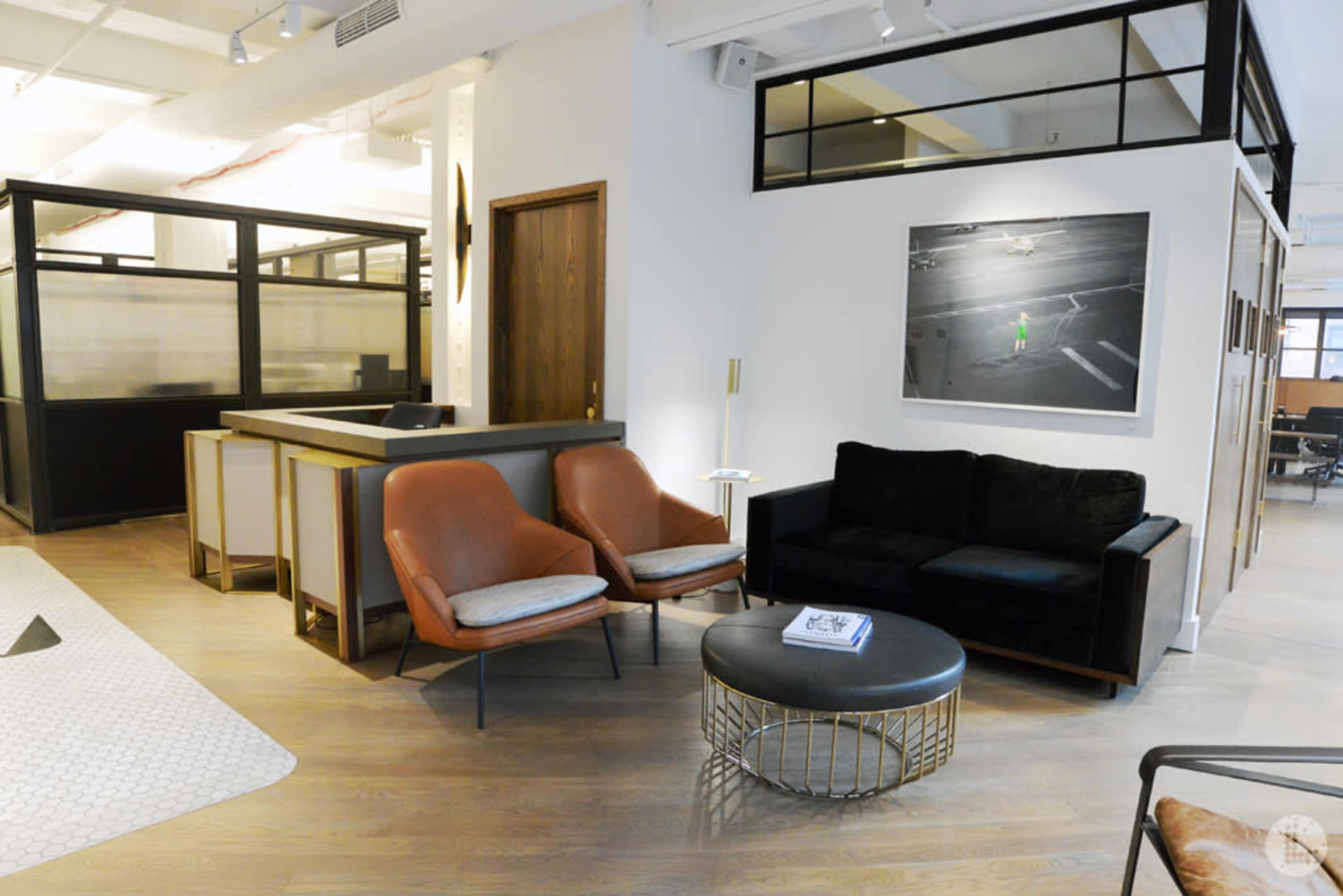 A modern office reception area with two orange chairs, a black couch, and a round coffee table, accompanied by a large artwork on the wall.