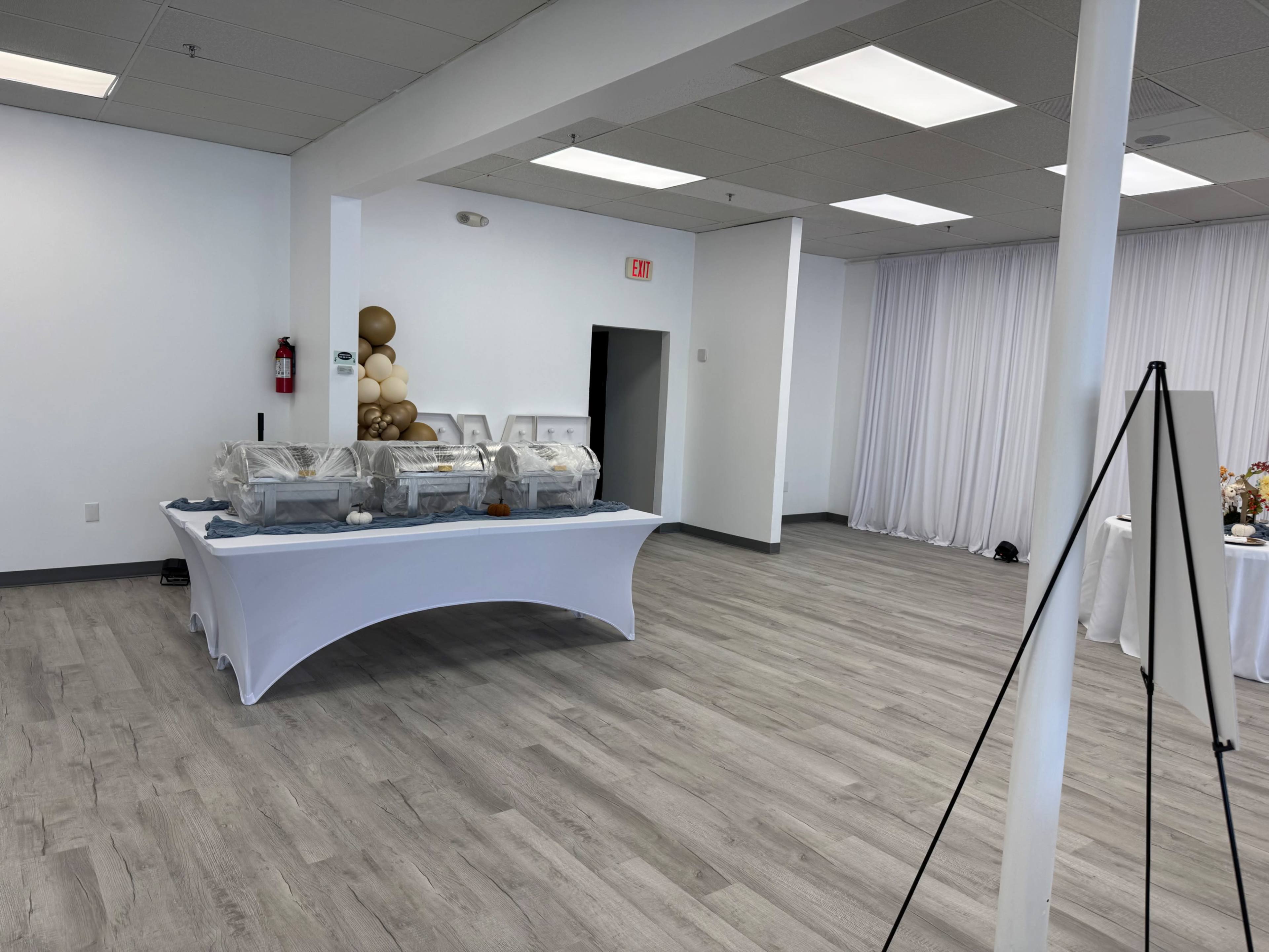 The image shows a spacious indoor venue with a buffet table covered by a white cloth, surrounded by plain walls and large windows, and a decorative arrangement of balloons in one corner.