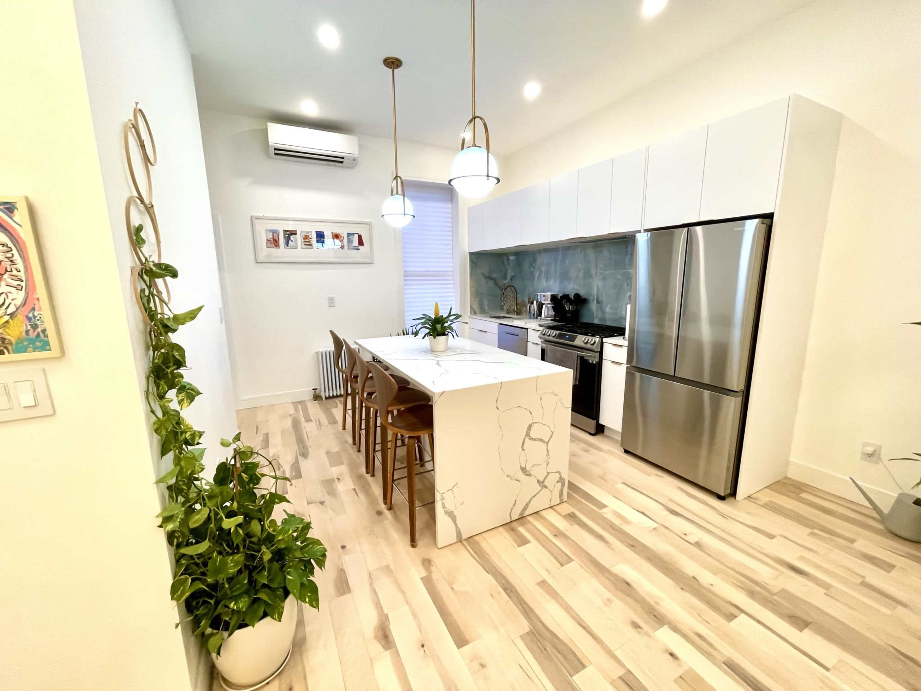 A modern kitchen features a marble-topped island with bar stools, stainless steel appliances, and light wood flooring.