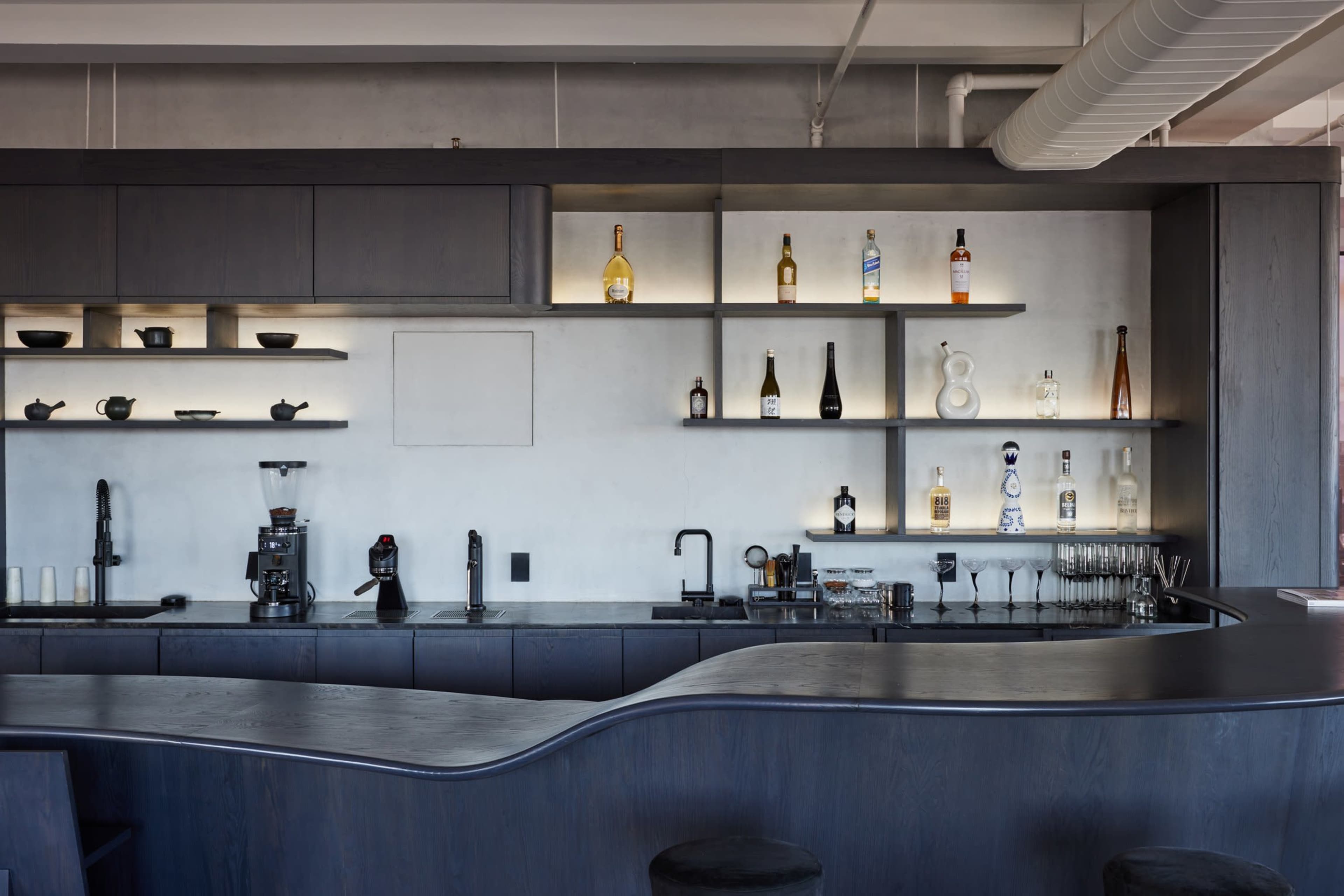 The image depicts a modern bar area with a sleek, curved countertop, shelves displaying various bottles, and minimalistic decor against a concrete wall.