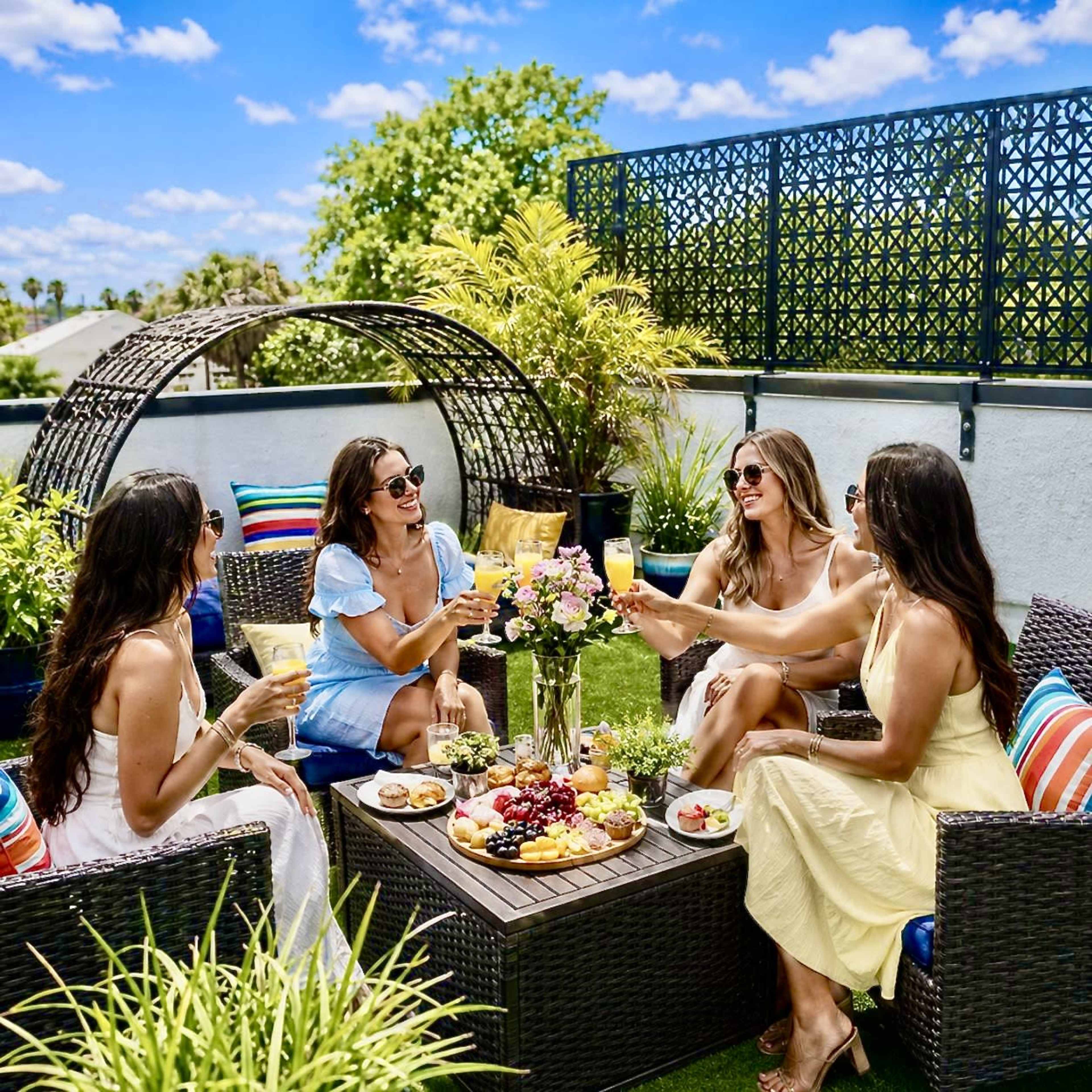 Five women are seated around a coffee table on a patio, enjoying drinks and a spread of fruits and snacks while surrounded by greenery under a blue sky.