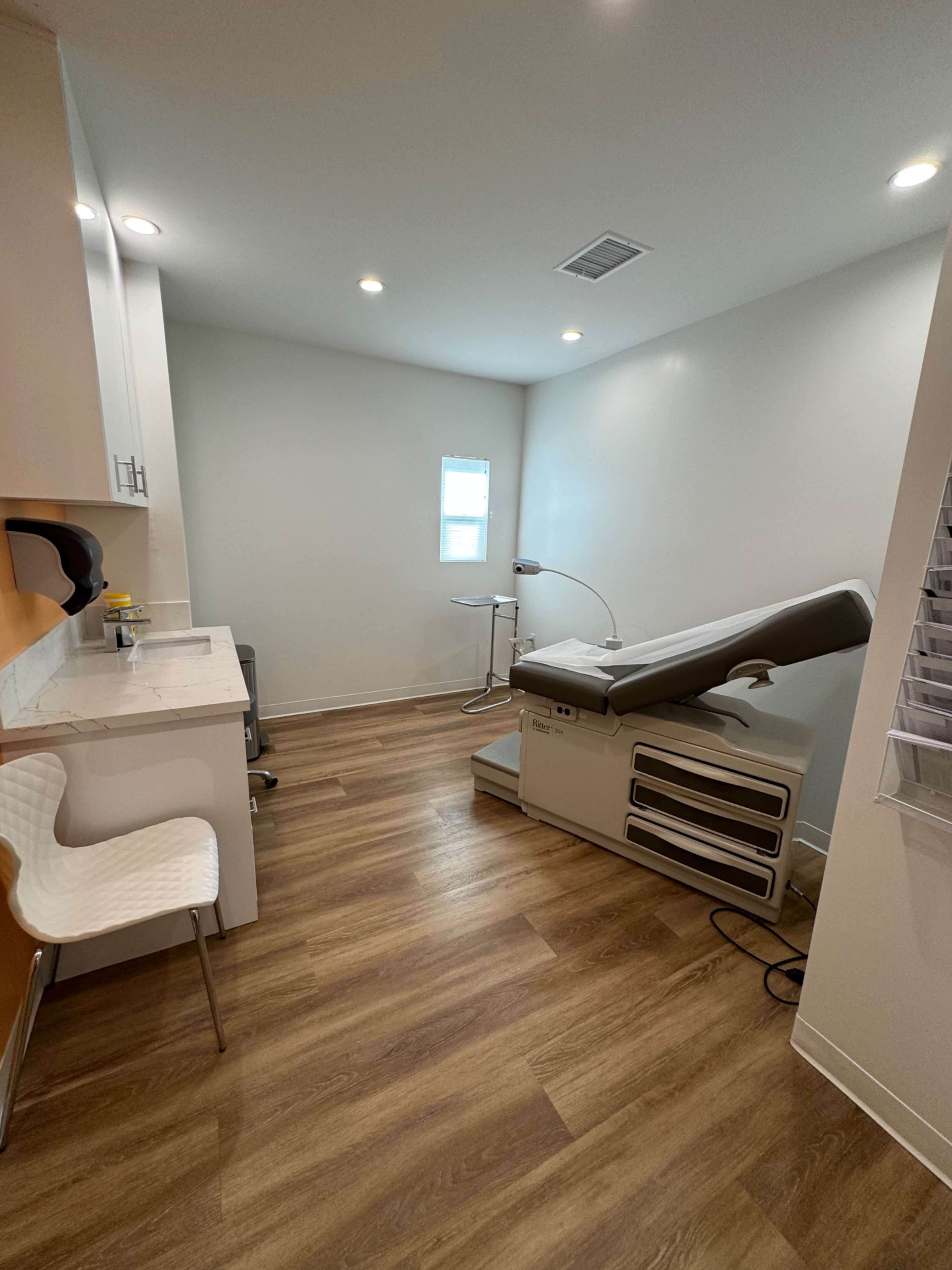 A medical examination room with a reclining examination table, a small desk, and wooden flooring.