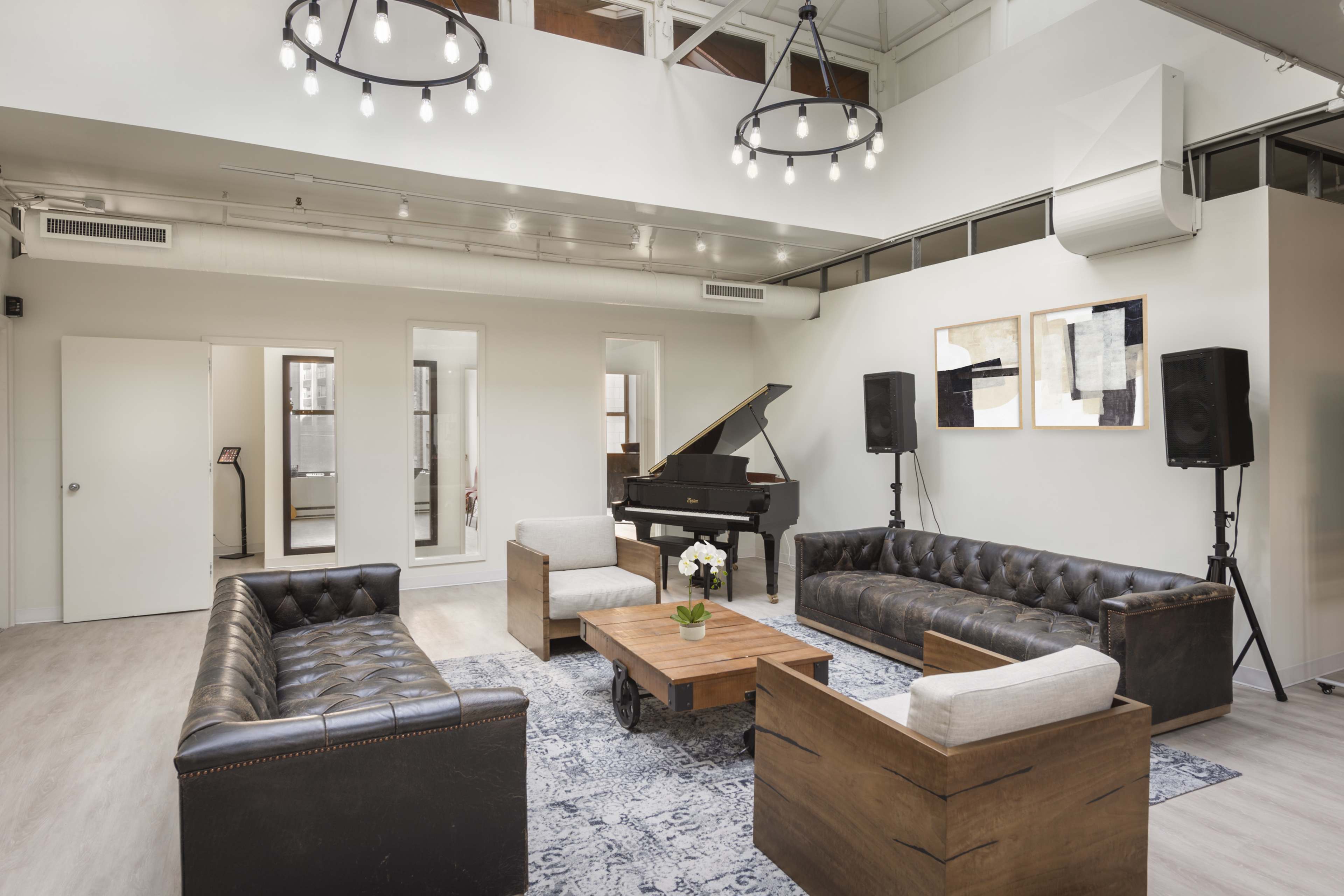 A modern lounge area with a grand piano, two leather sofas, a wooden coffee table with a vase of flowers, and large mirrors on the walls.