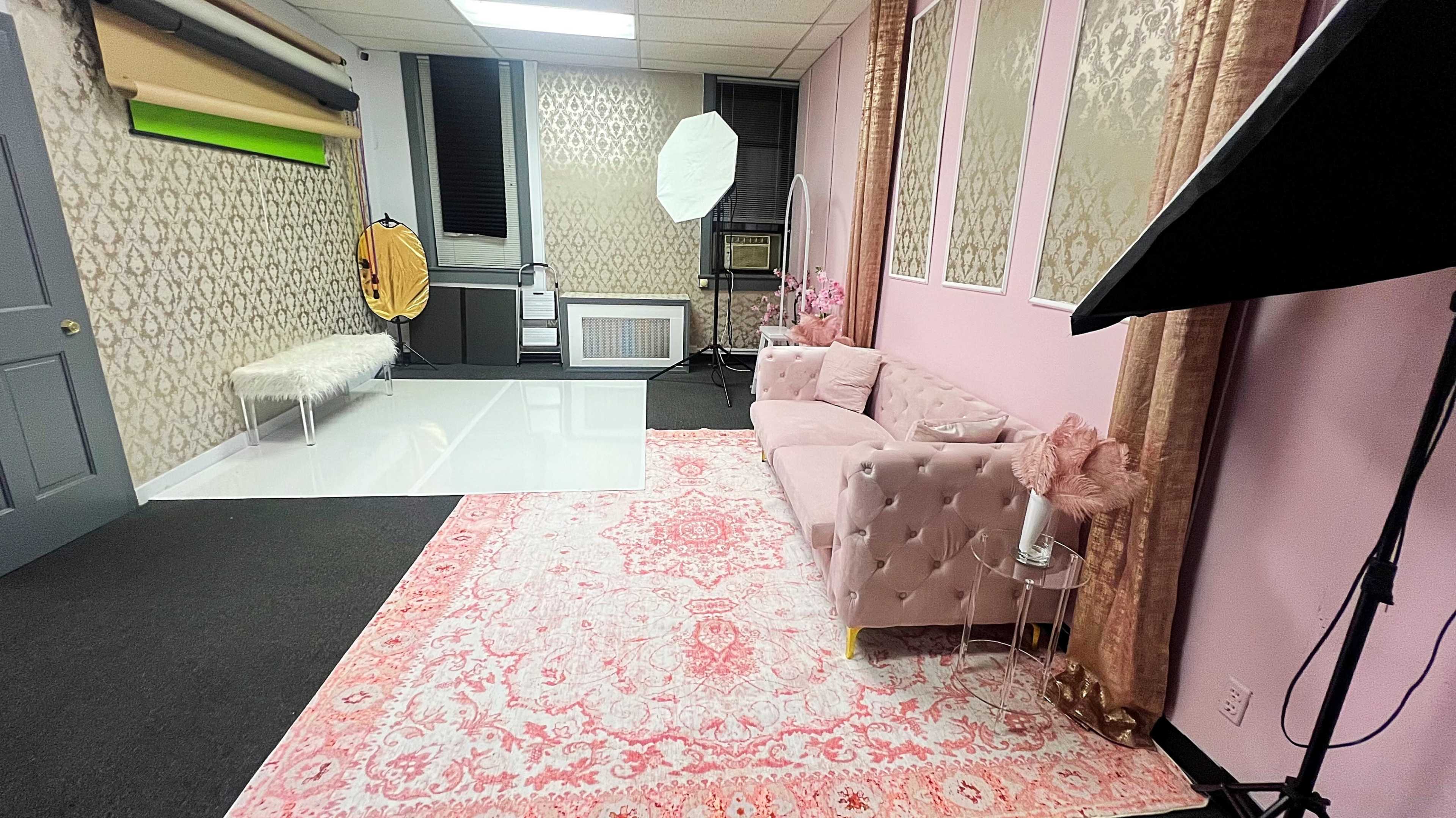 The image shows a styled photography studio with a pink couch, a large white backdrop area, and various lighting equipment arranged around a patterned rug.