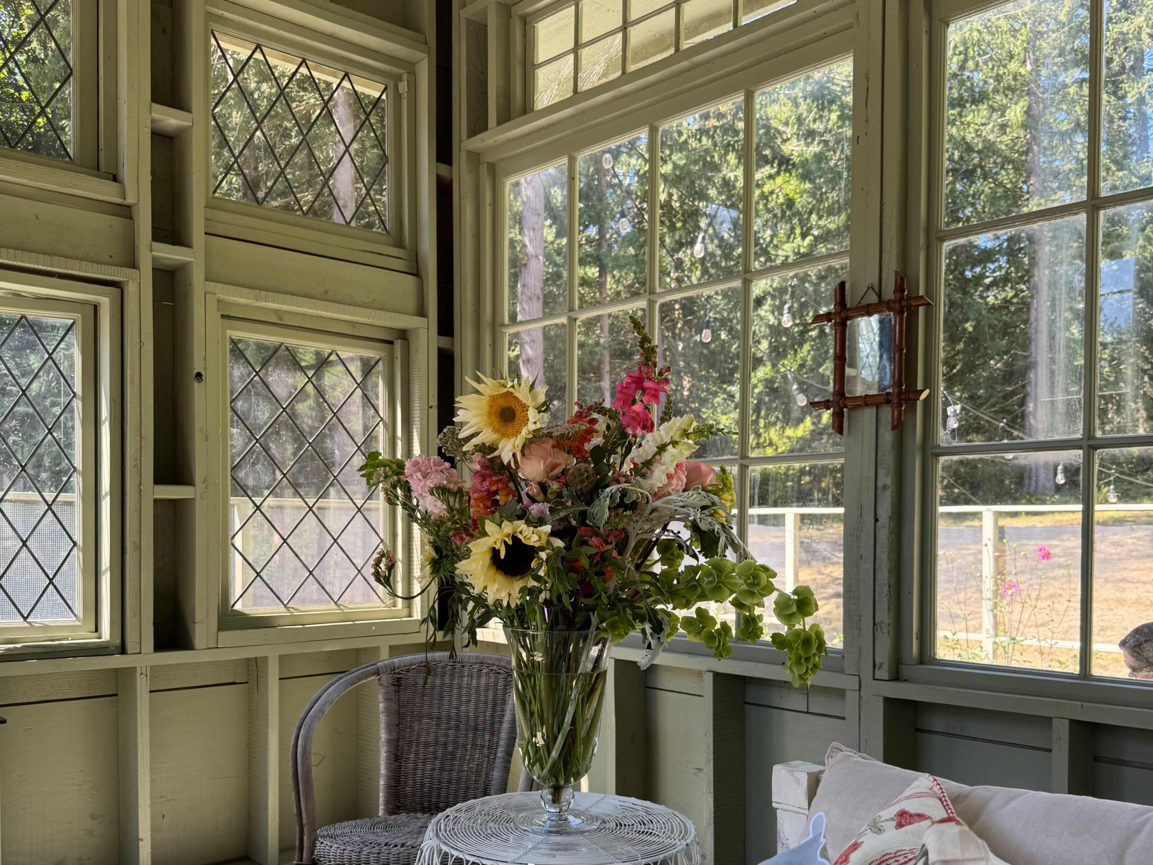 A vase of mixed flowers sits on a small table inside a sunlit room with large windows and green walls.