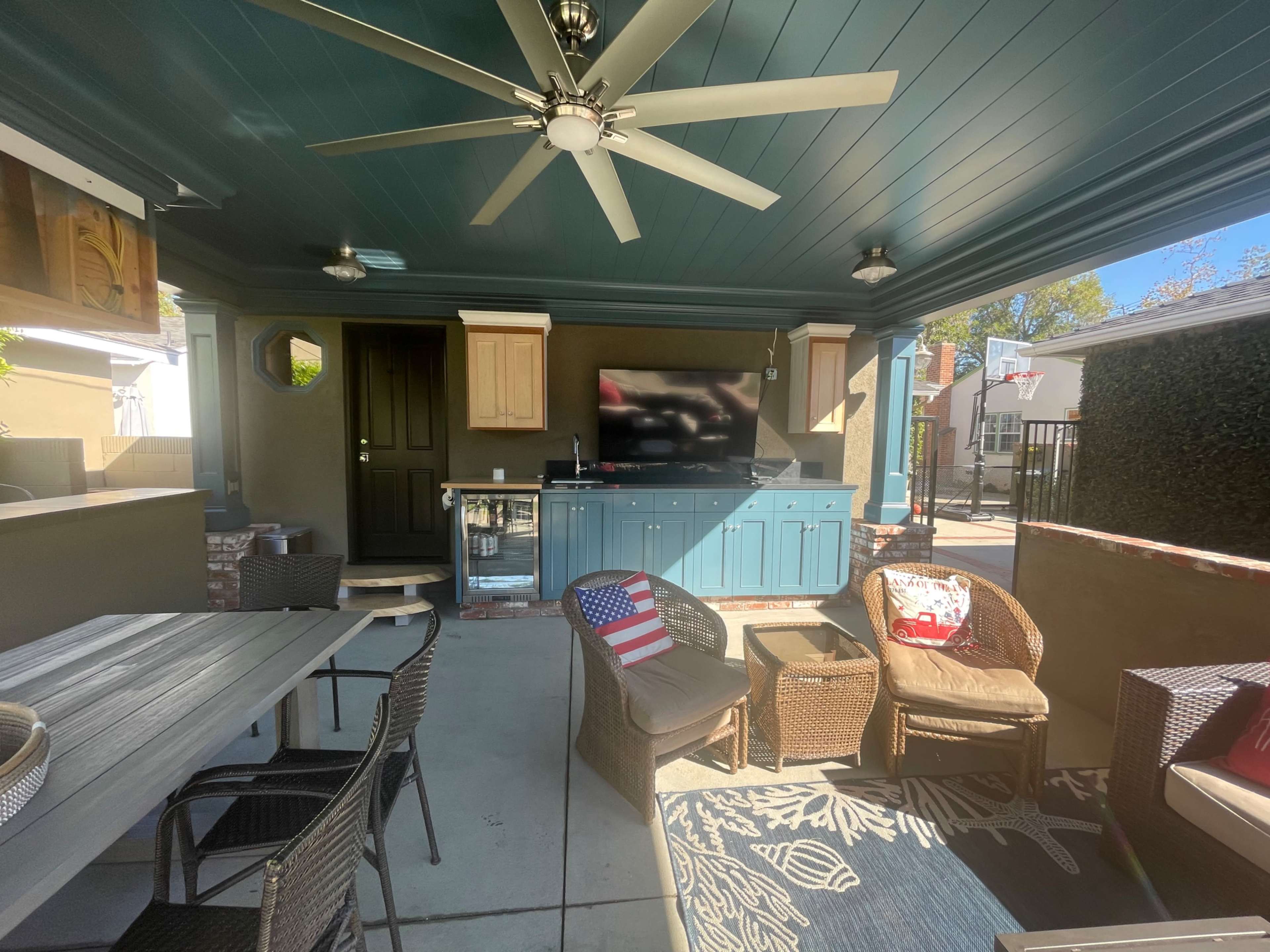 The image shows a covered outdoor patio featuring a dining table, wicker chairs, a television mounted on the wall, and a kitchenette area.