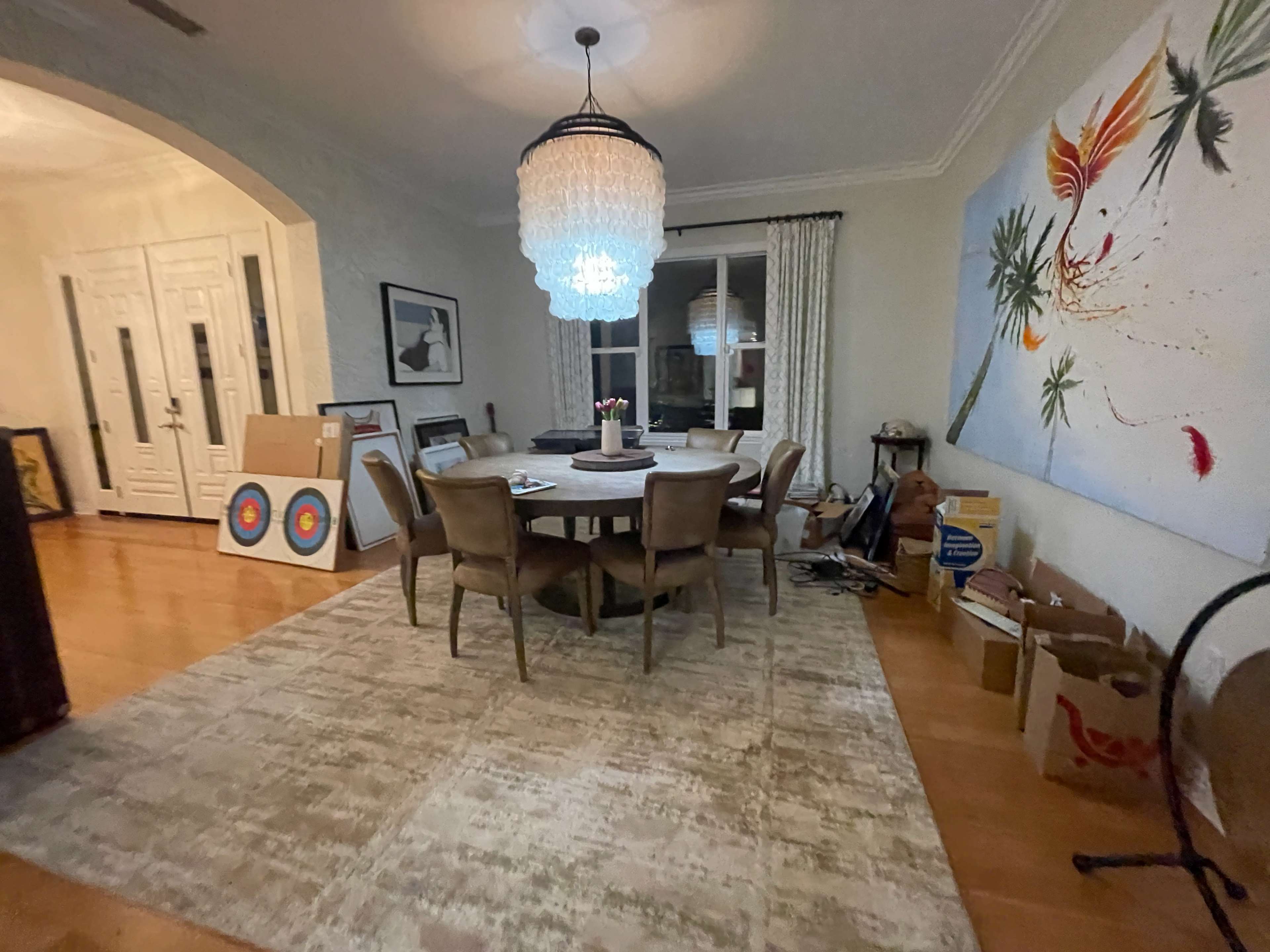 A dining room features a round table surrounded by chairs, with a large chandelier above and various artwork and boxes placed around the space.