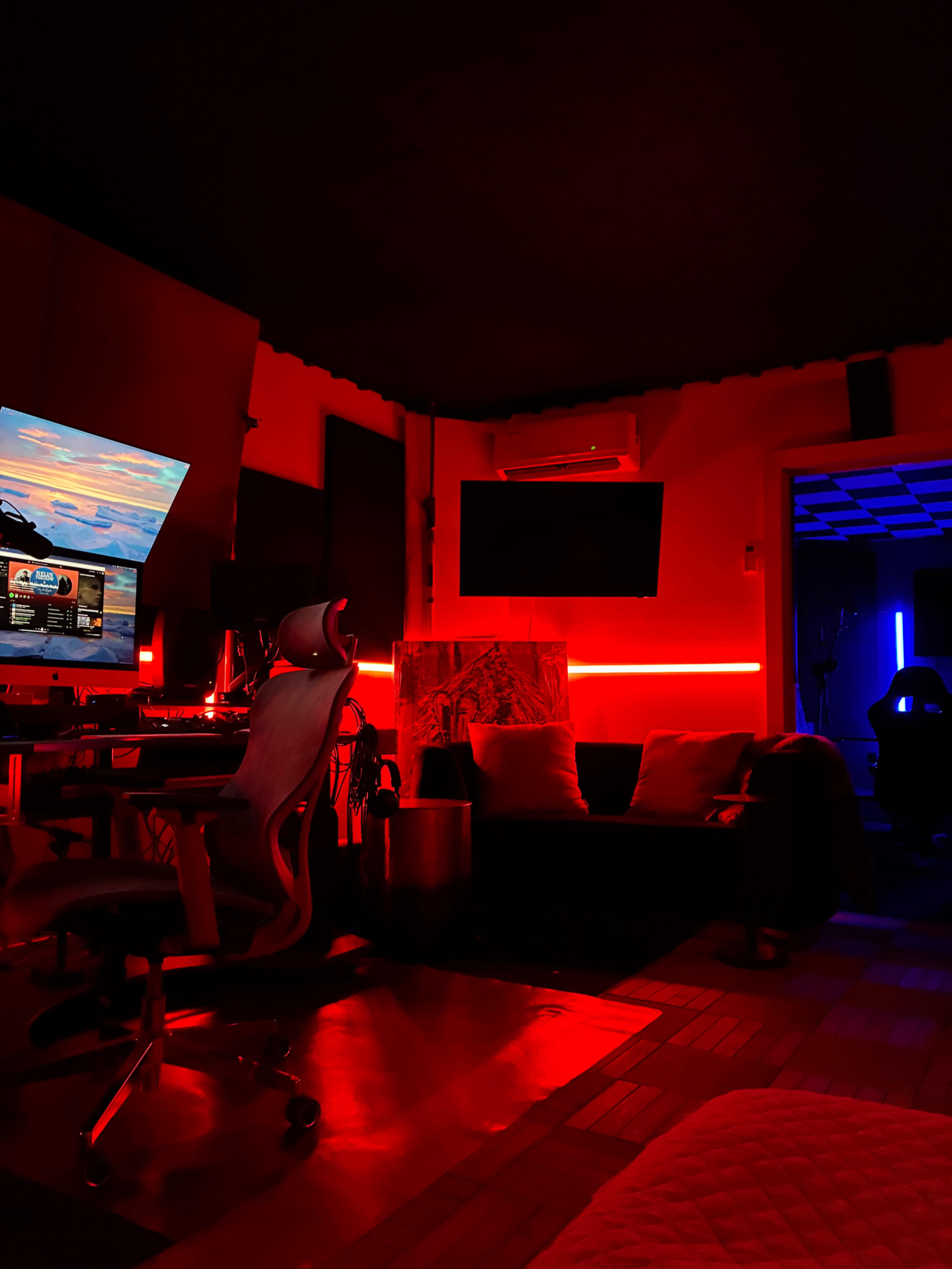 A dimly lit room features a gaming setup with a large monitor, red ambient lighting, and a cozy seating area.