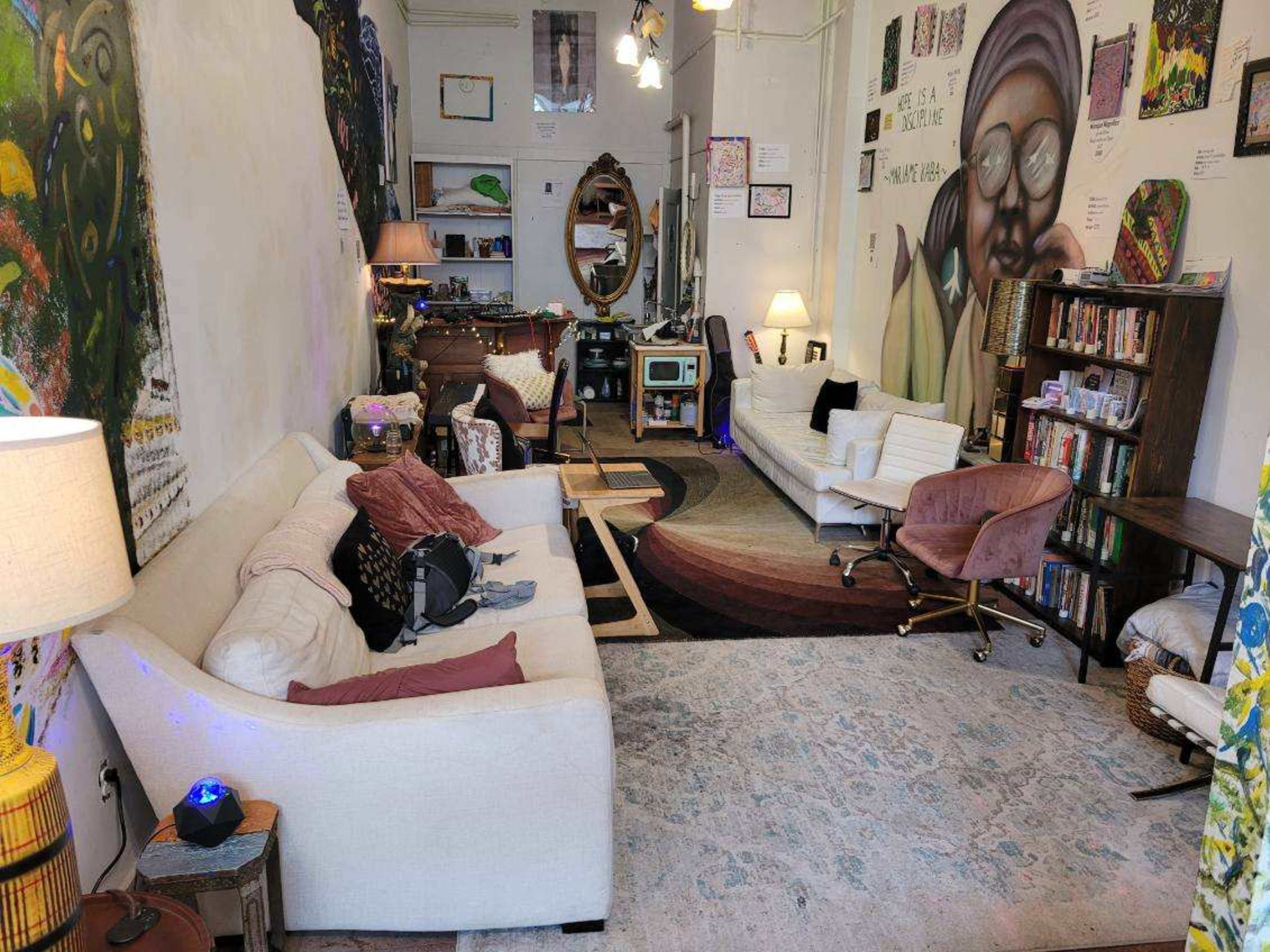 The image shows a cozy living space with a mix of seating, a large mirror, bookshelves, and vibrant wall art.