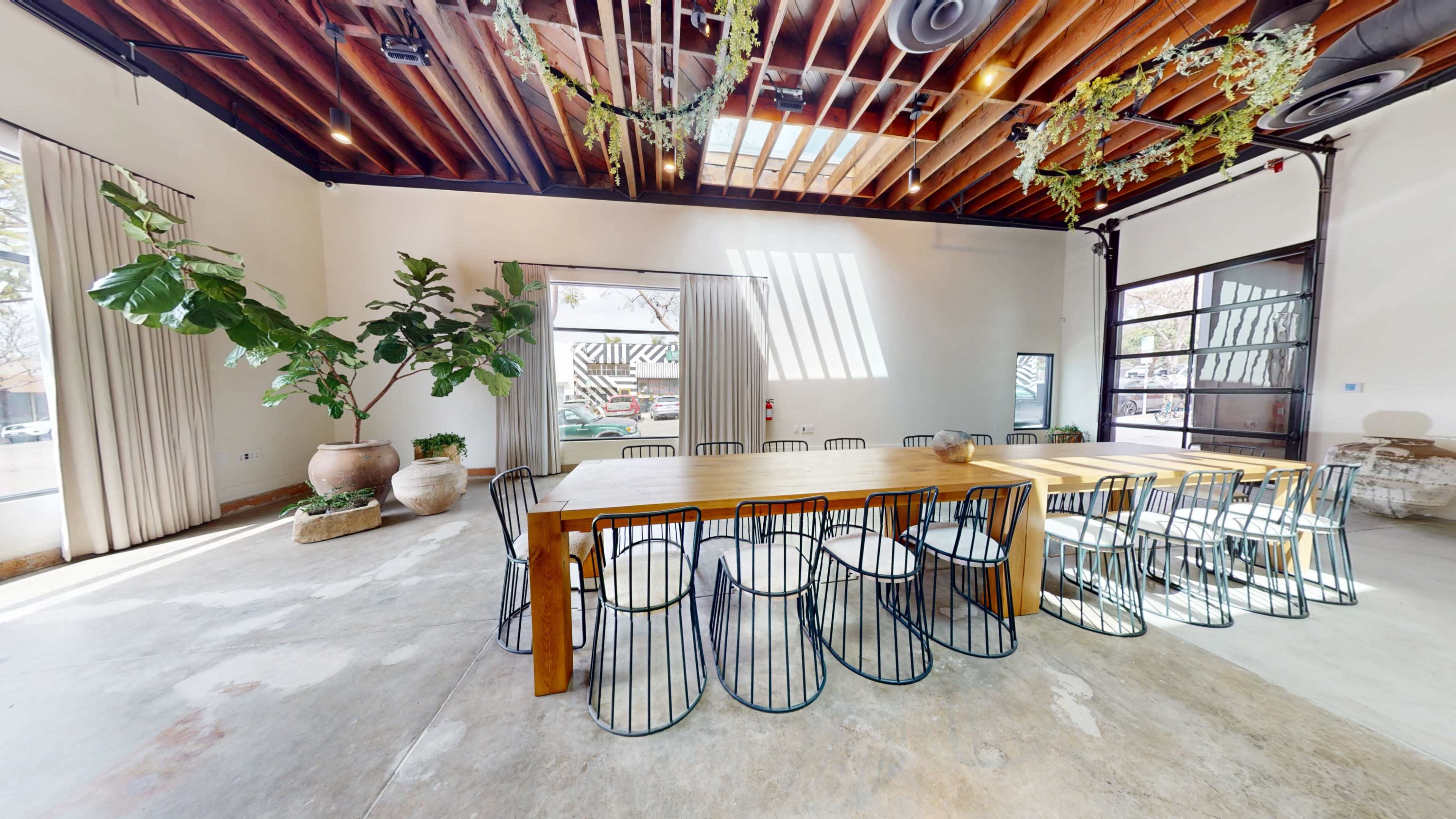 The image shows a spacious room featuring a long wooden table surrounded by wire chairs, with large windows and potted plants adding natural elements.