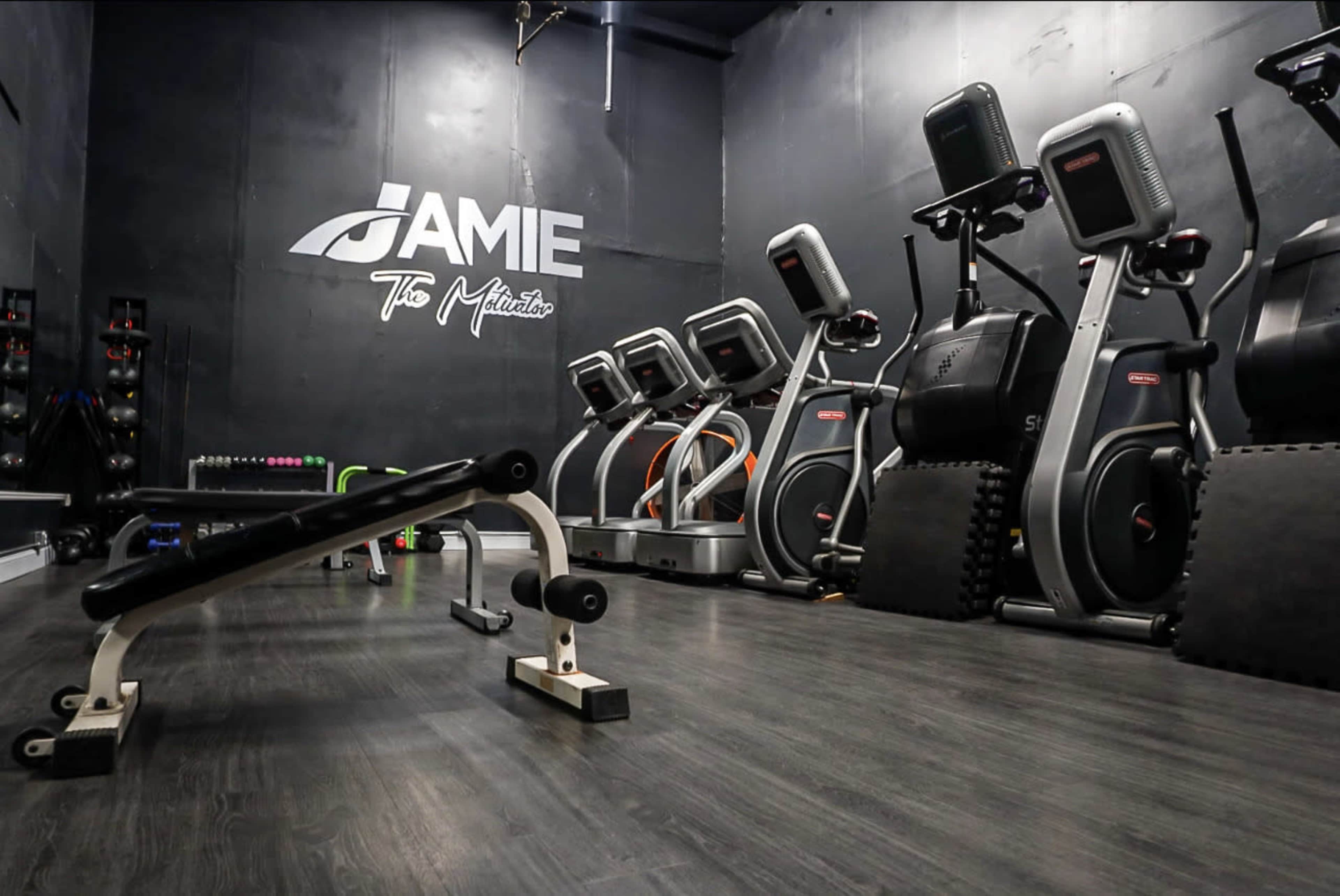 A fitness studio with exercise equipment, including elliptical machines and a weight bench, set against a black wall with the name "Jamie" displayed prominently.