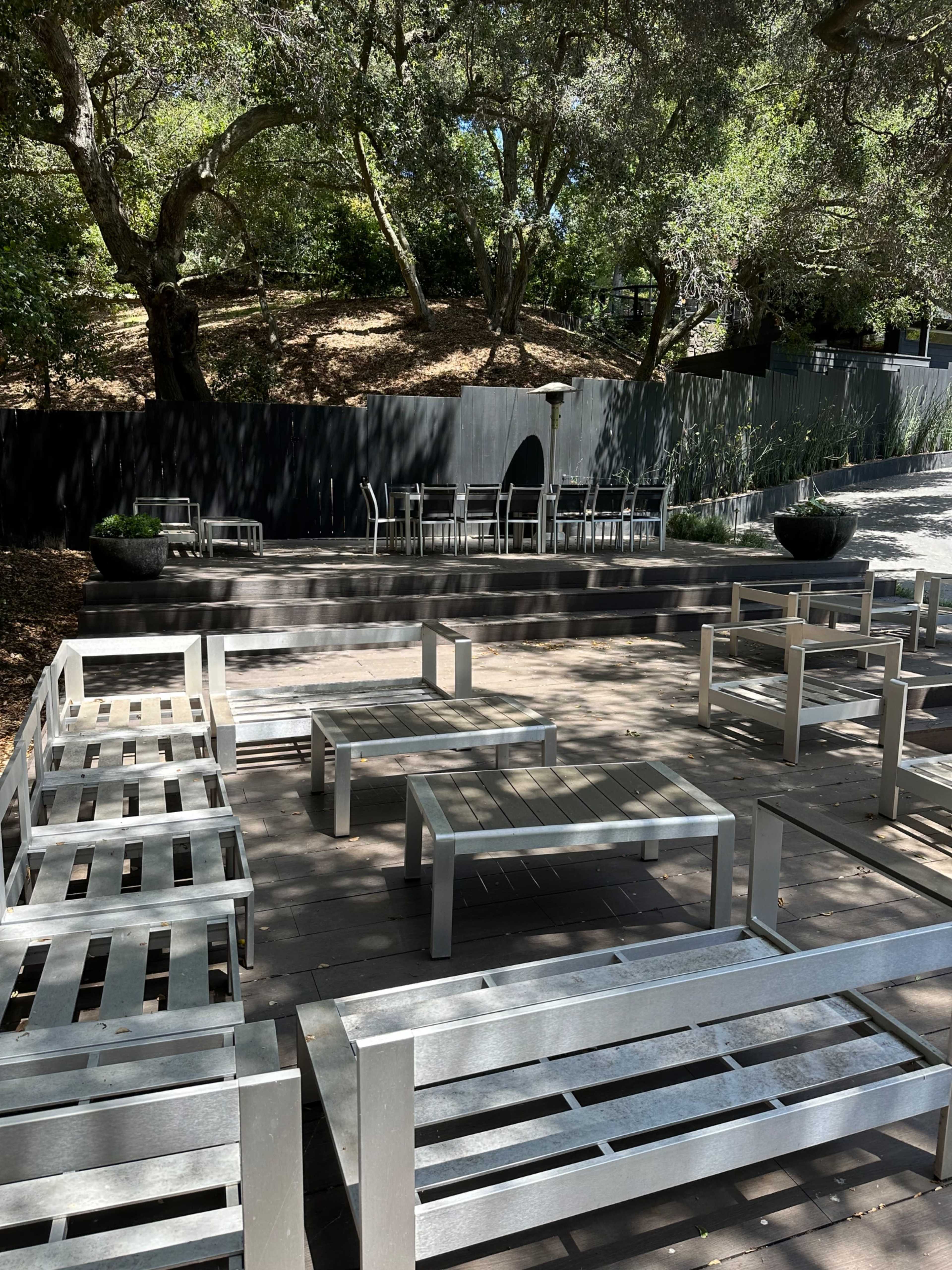 A series of metal outdoor furniture arranged on a paved area under trees, with additional seating visible in the background.