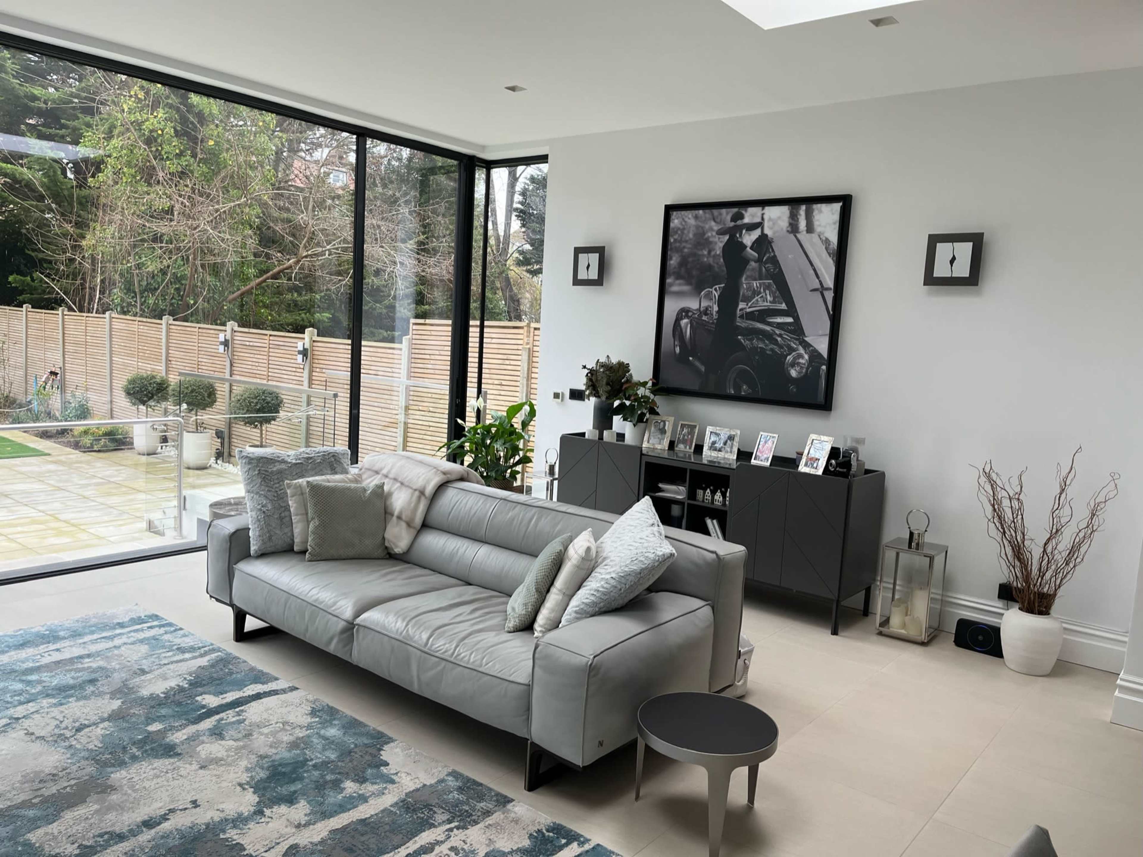 A modern living room with a gray leather sofa, a large black-and-white photograph on the wall, and large windows overlooking a garden.