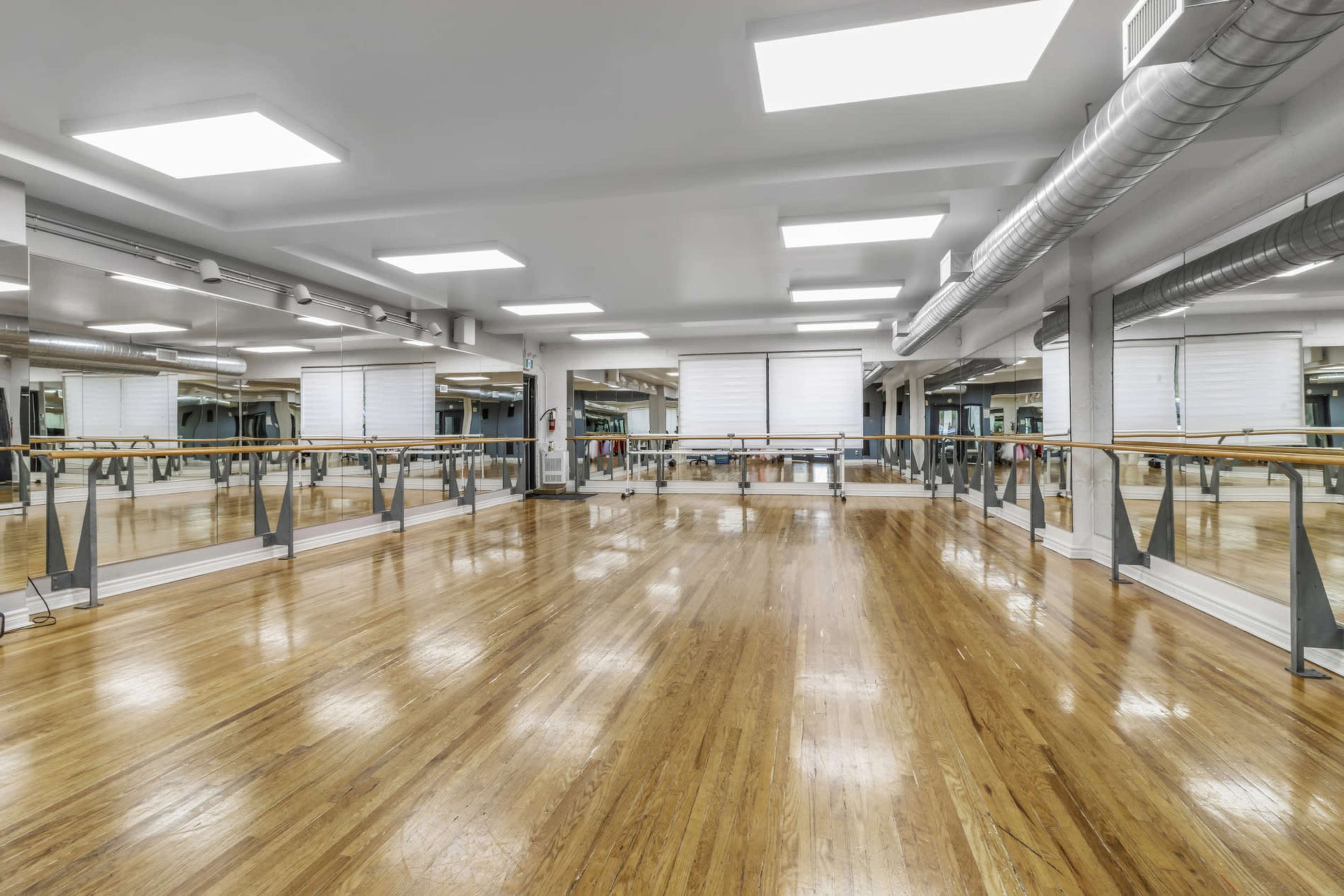 The image shows a spacious dance studio with wooden floors, large mirrors, and bright overhead lights.